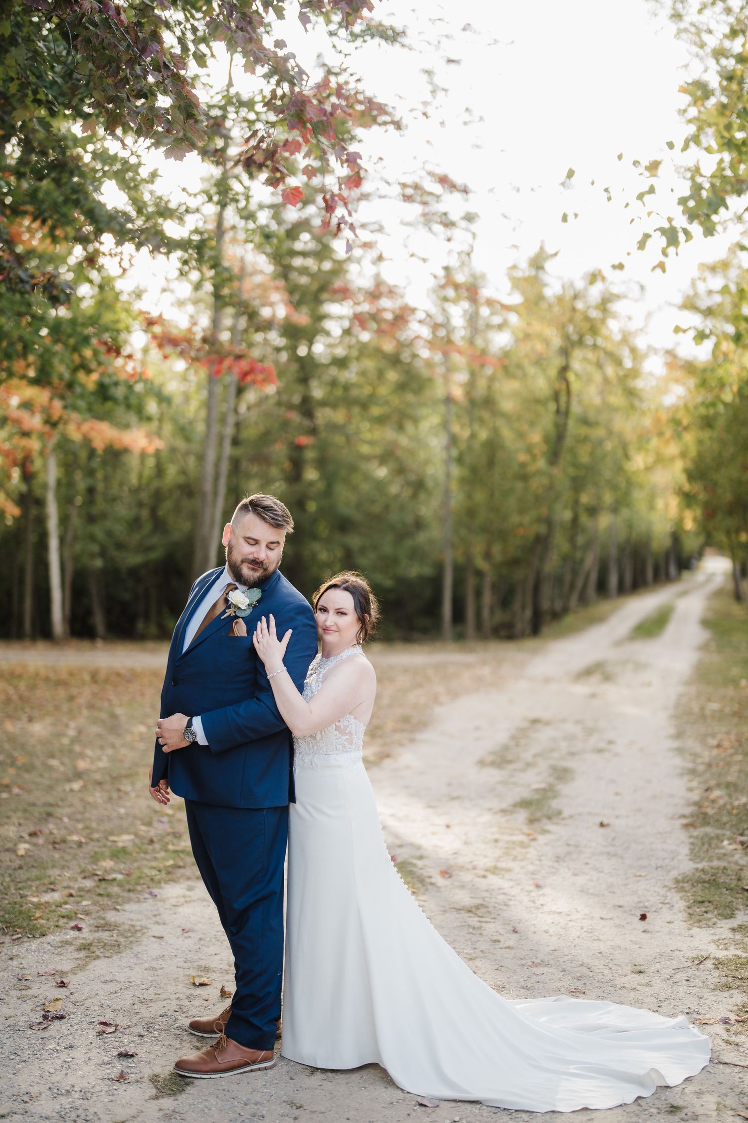 A joyful wedding couple poses on a tree-lined autumn path in formal attire.