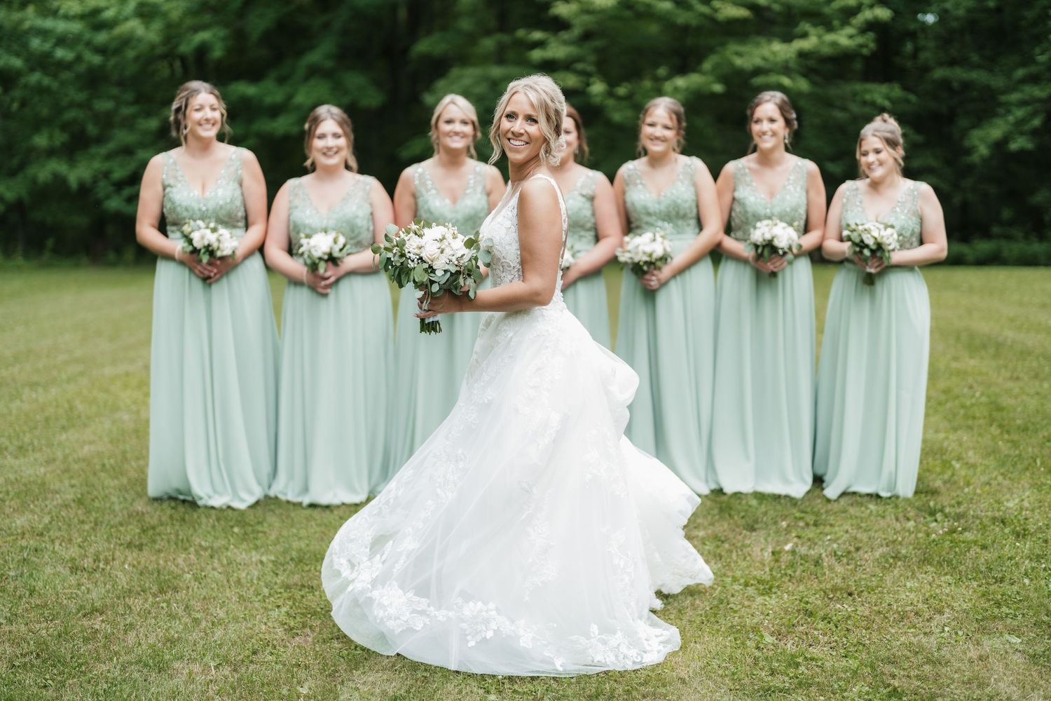Wedding party poses on green lawn with bridesmaids in soft mint dresses and bride in white gown.
