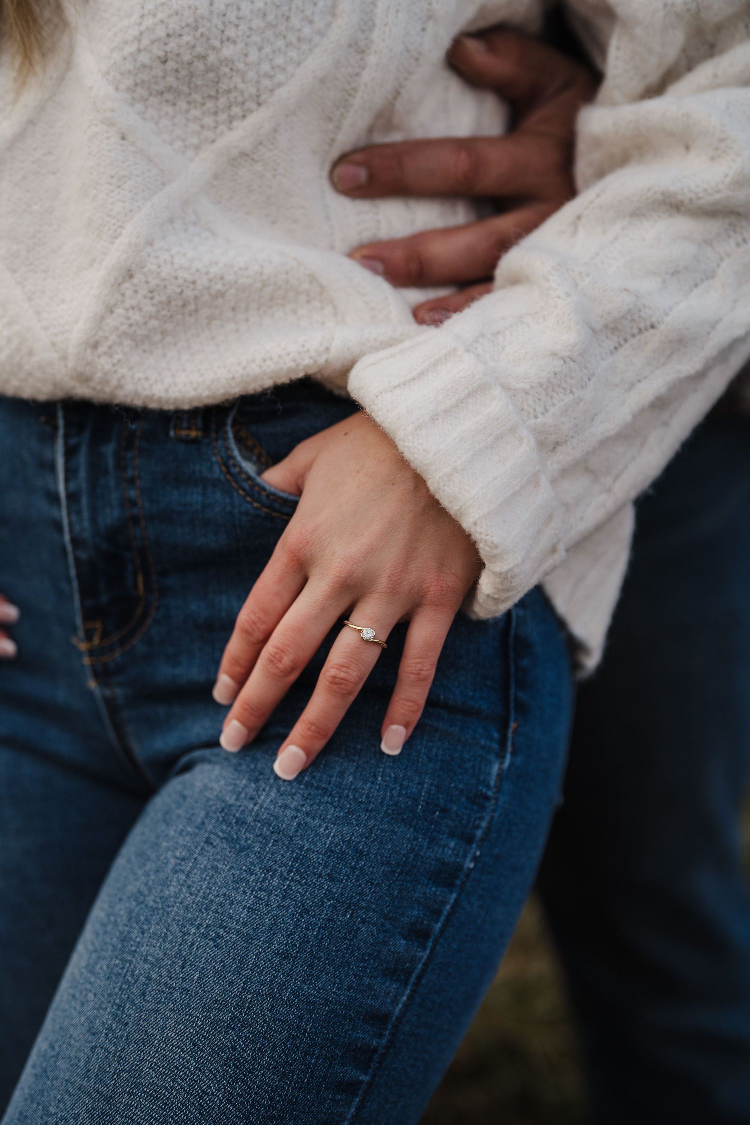 Close-up of hands wearing a white sweater and blue jeans, showing an engagement ring.
