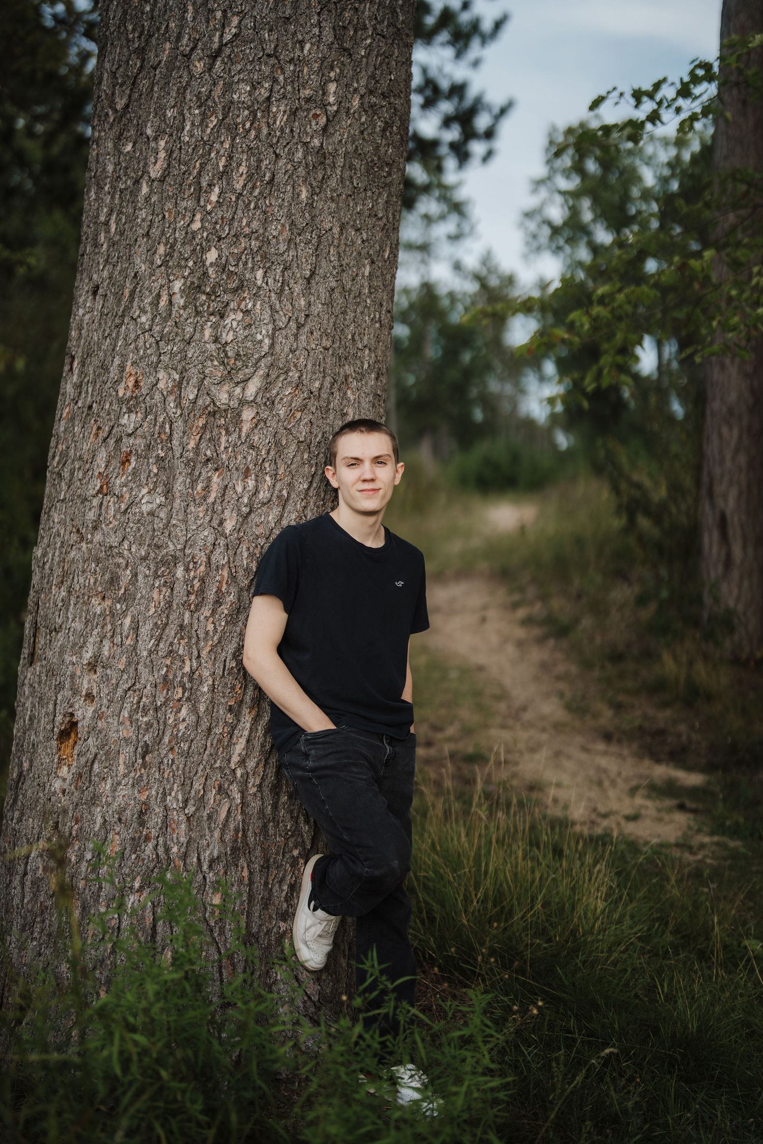 A casual portrait against a large tree trunk on a wooded path wearing a black polo shirt and dark pants.