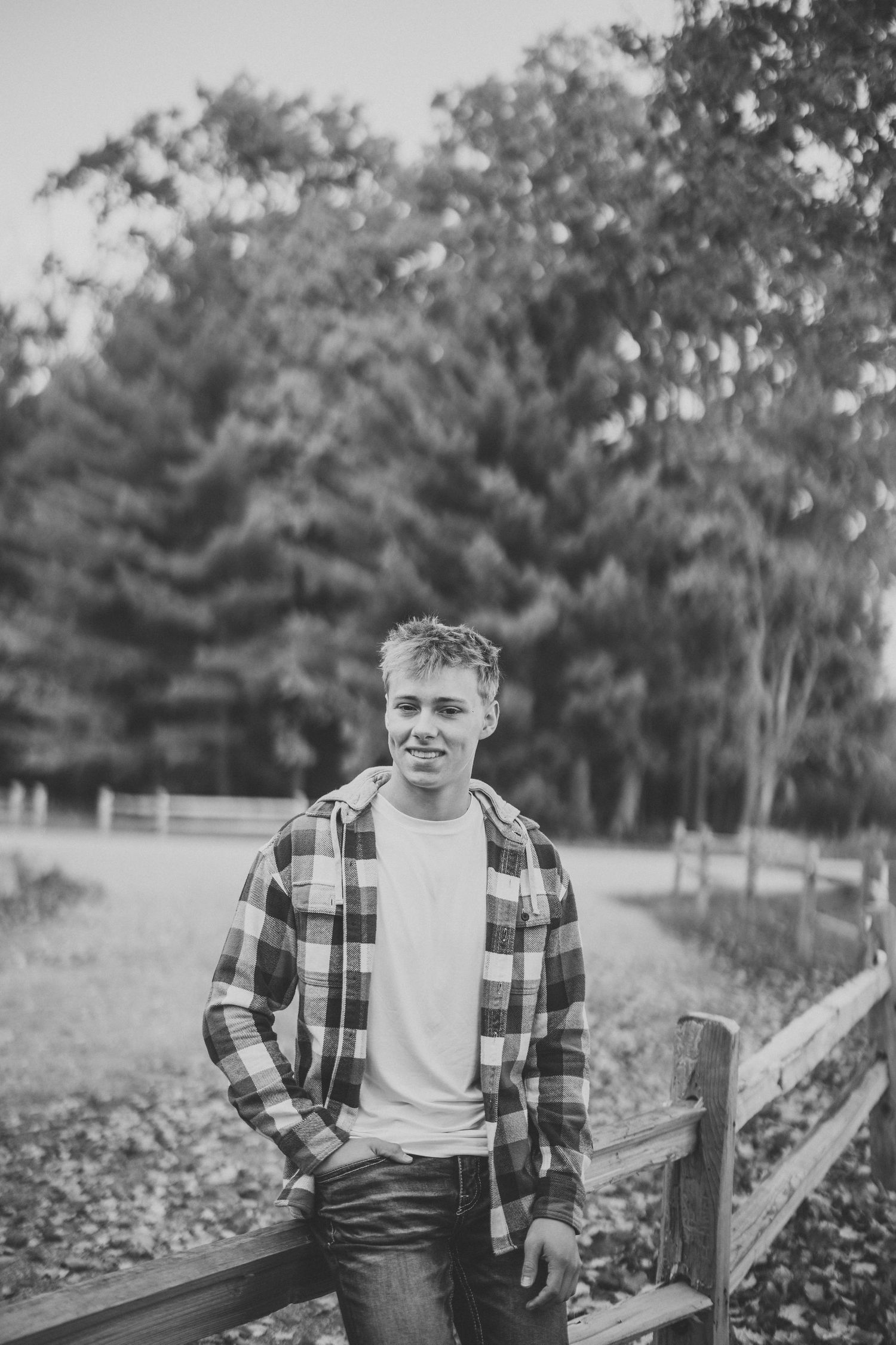 Black and white portrait of someone in a plaid flannel shirt leaning against a wooden fence in a rural setting.