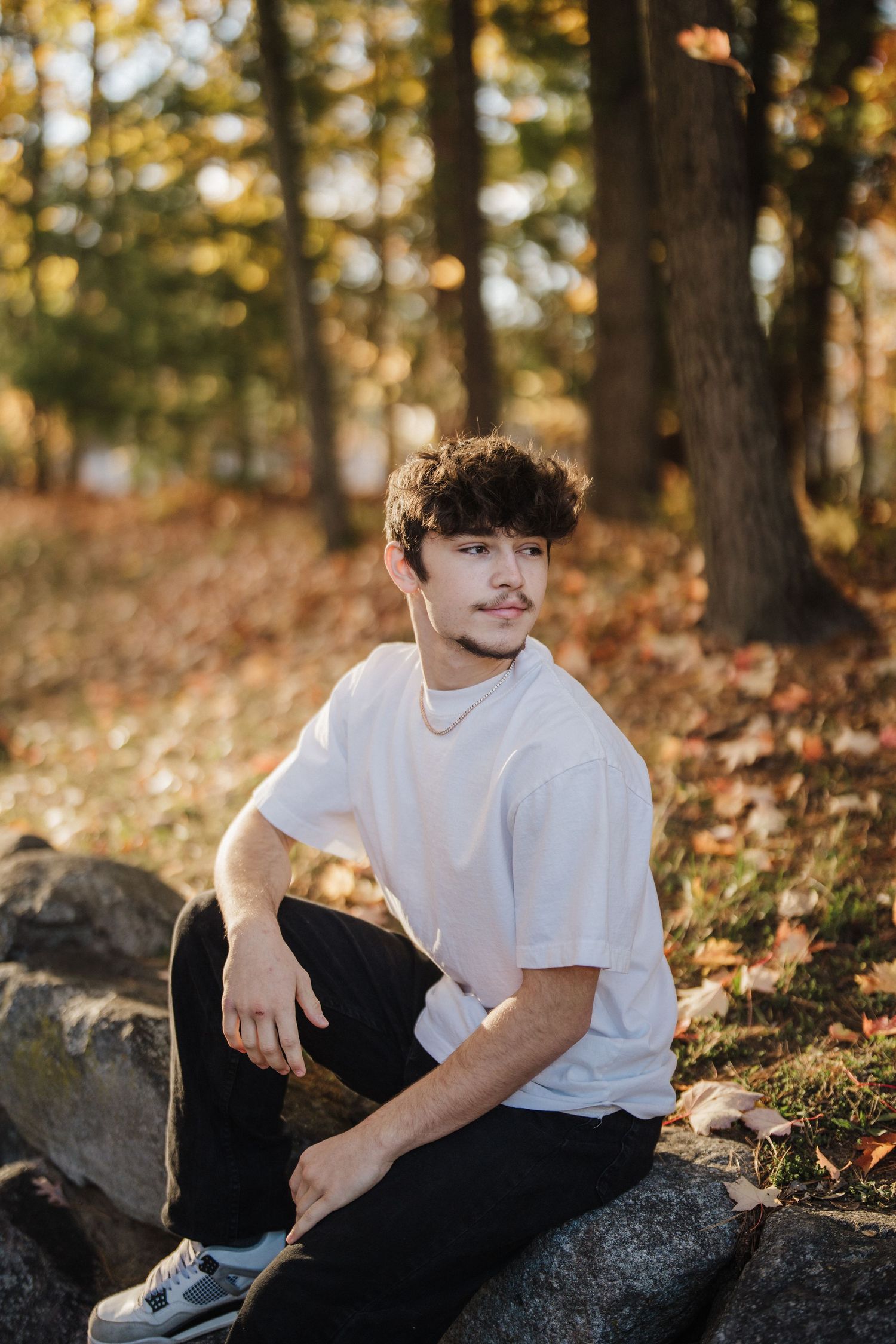 A person in a white t-shirt and dark pants sits casually on rocks surrounded by autumn foliage in a forest setting.