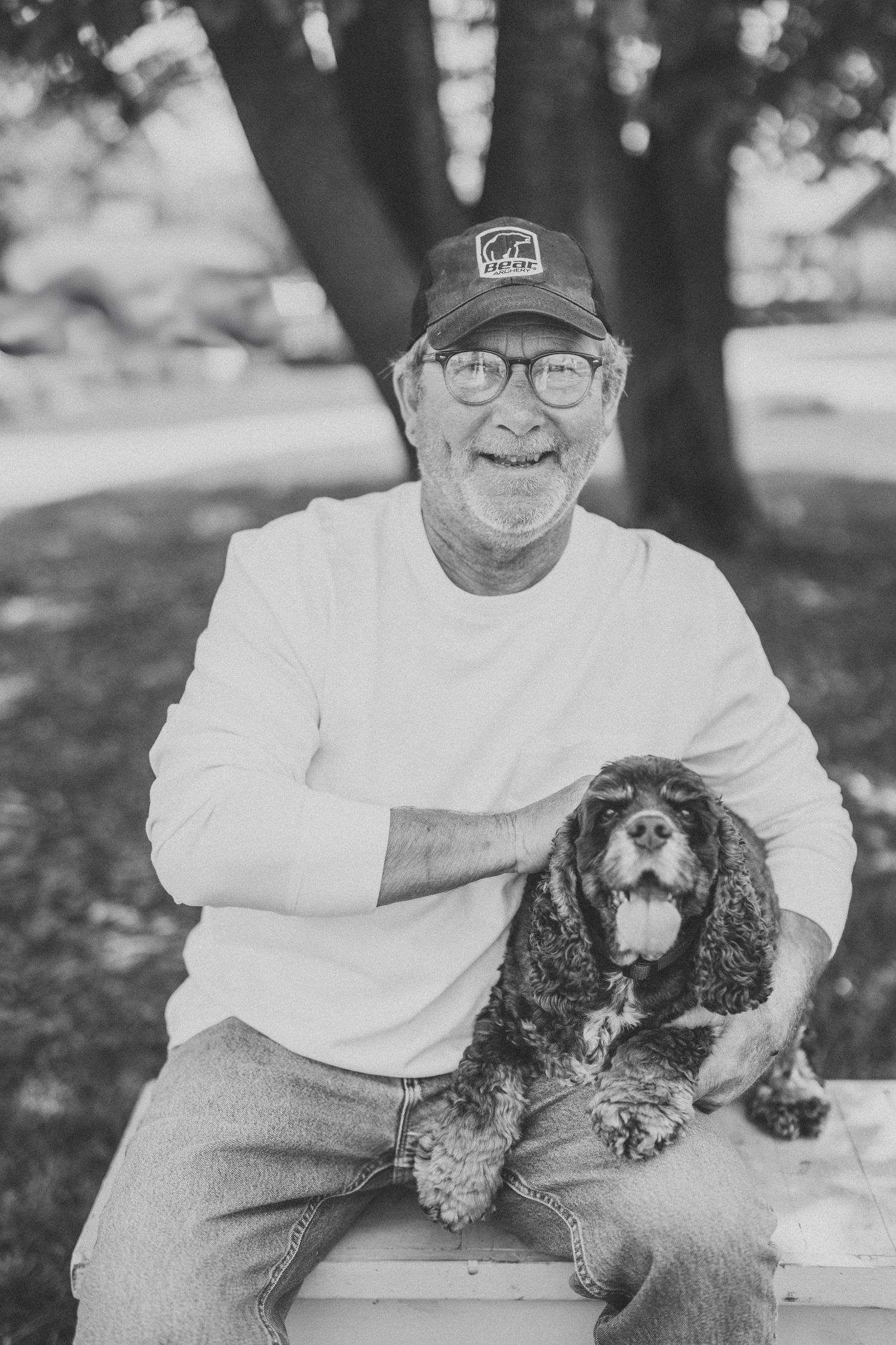 A black and white photo of someone sitting with a small spaniel dog on their lap in an outdoor setting.