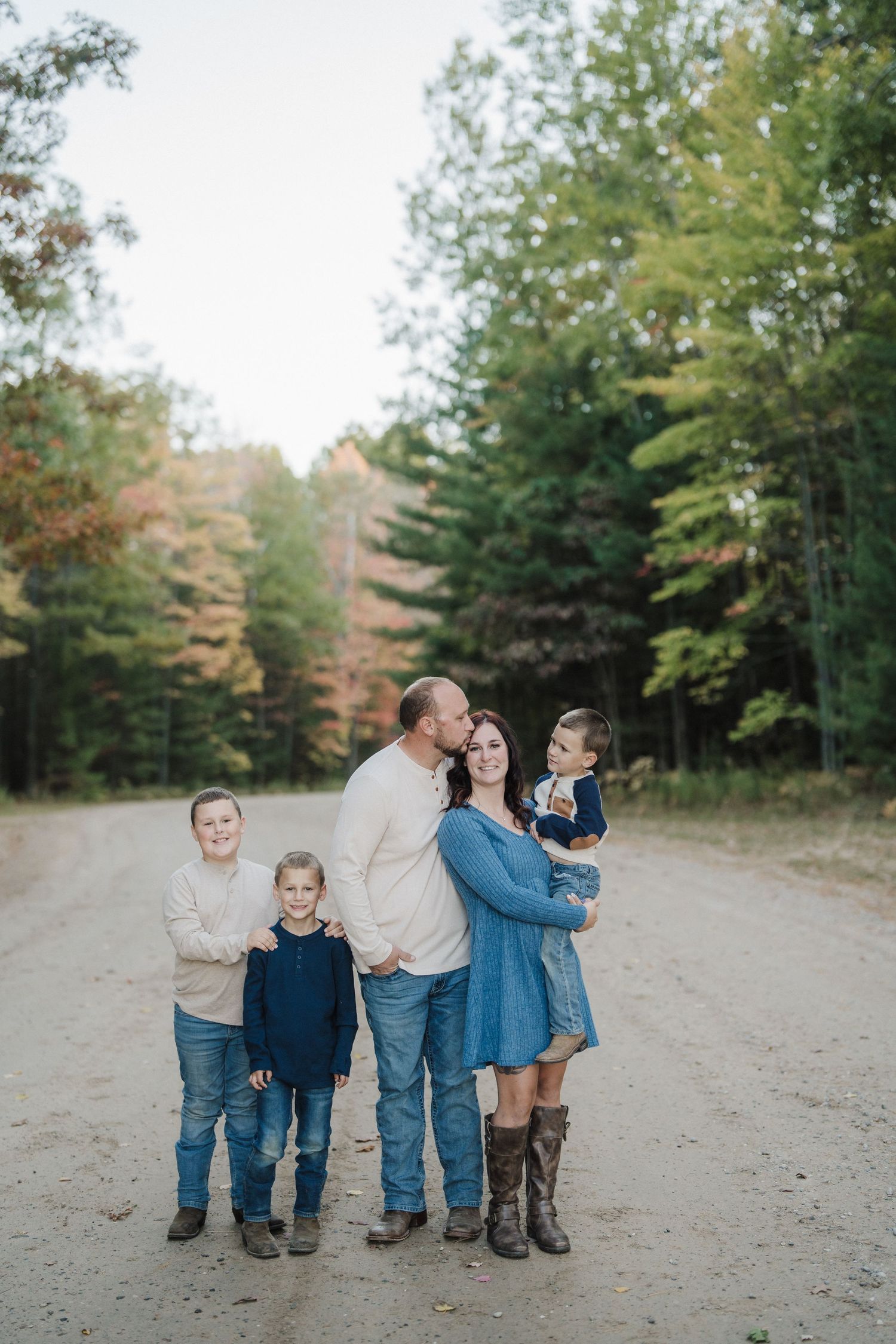 A group in coordinating denim and neutral outfits stands together on a dirt path surrounded by autumn trees.