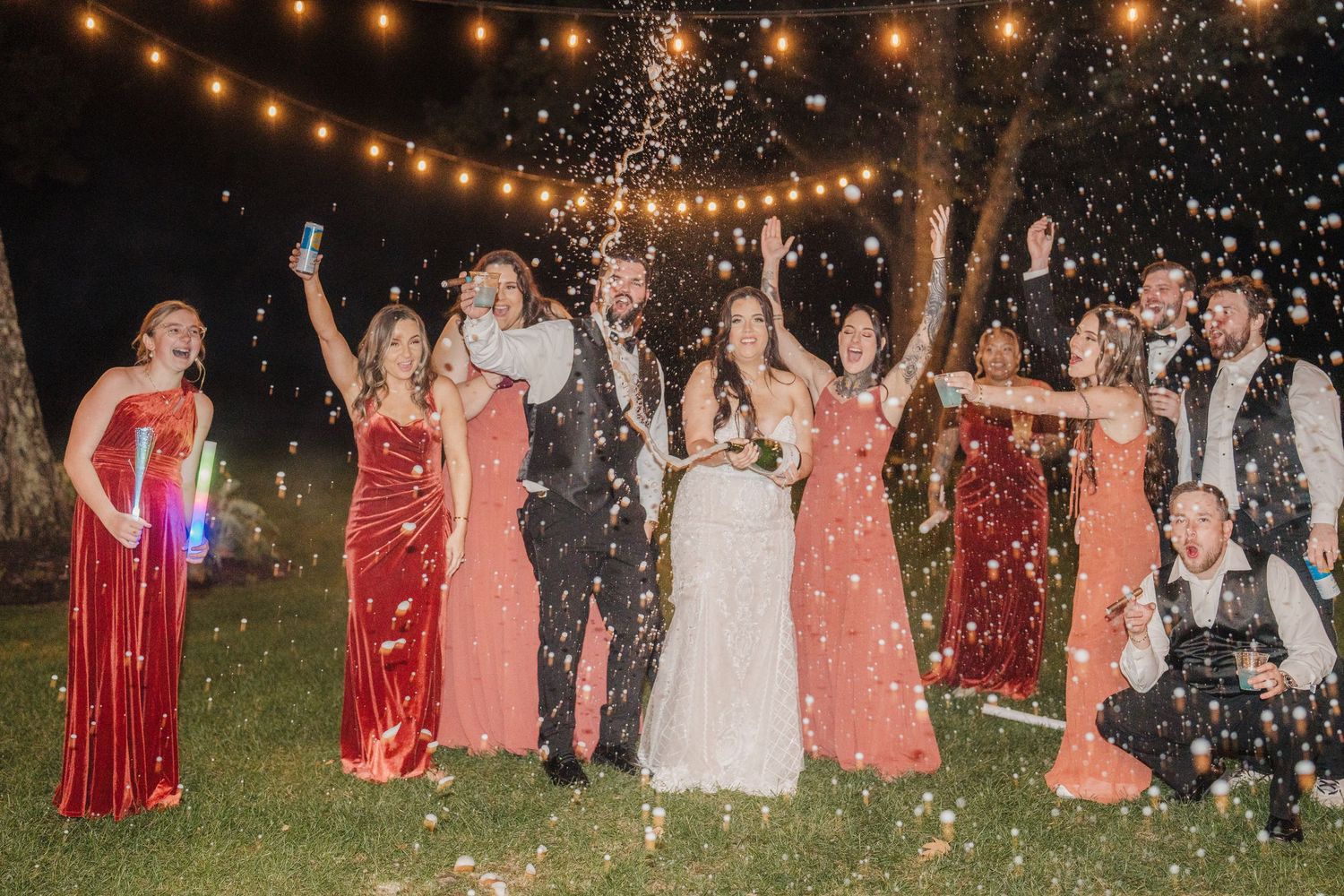 Wedding party celebrates under string lights with sparklers creating magical nighttime atmosphere.