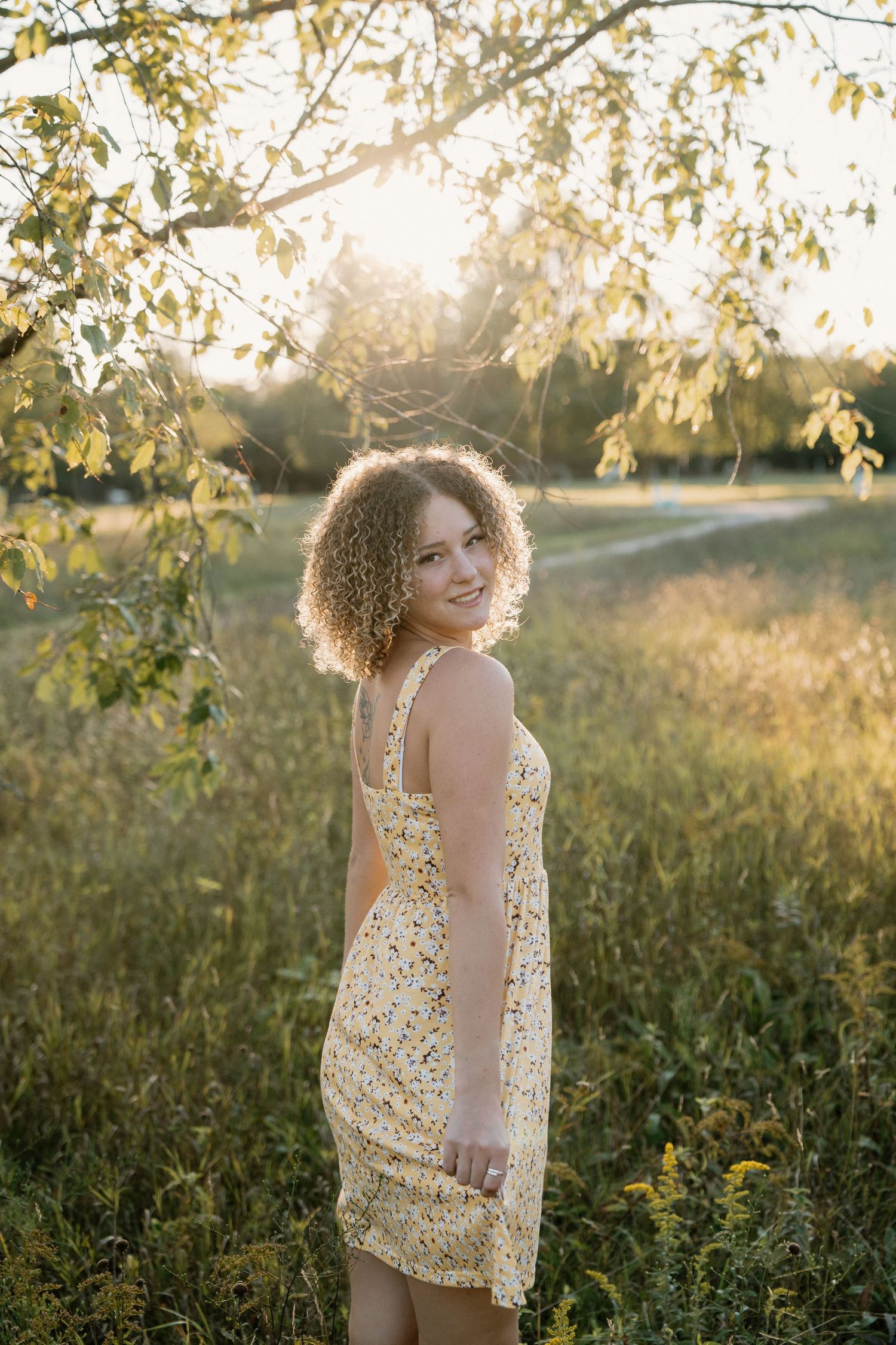 Young woman with curly hair in floral sundress smiling outdoors in golden sunset light among green fields.