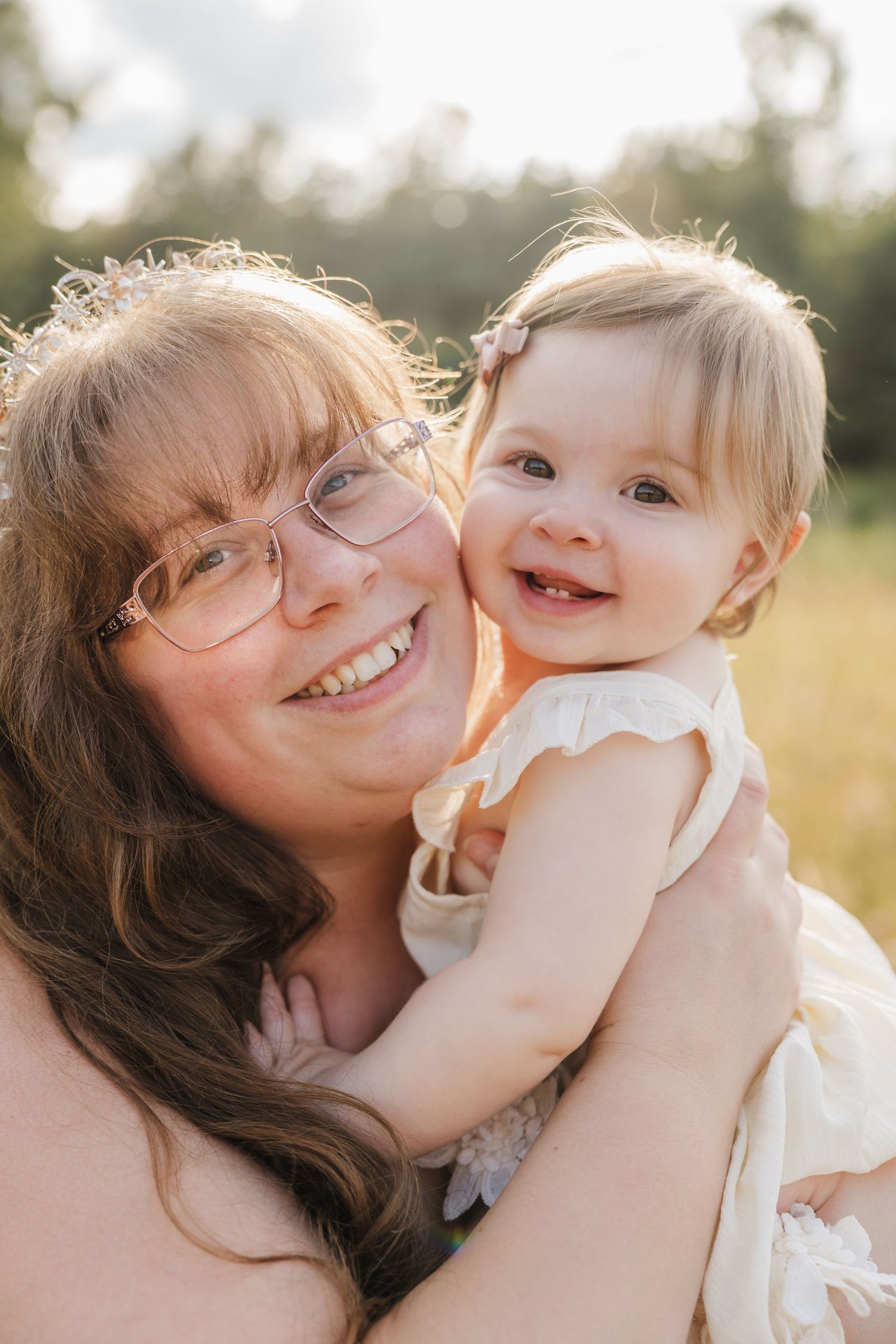 A smiling woman with glasses holds a happy baby girl in a white dress outdoors in a sunny field.