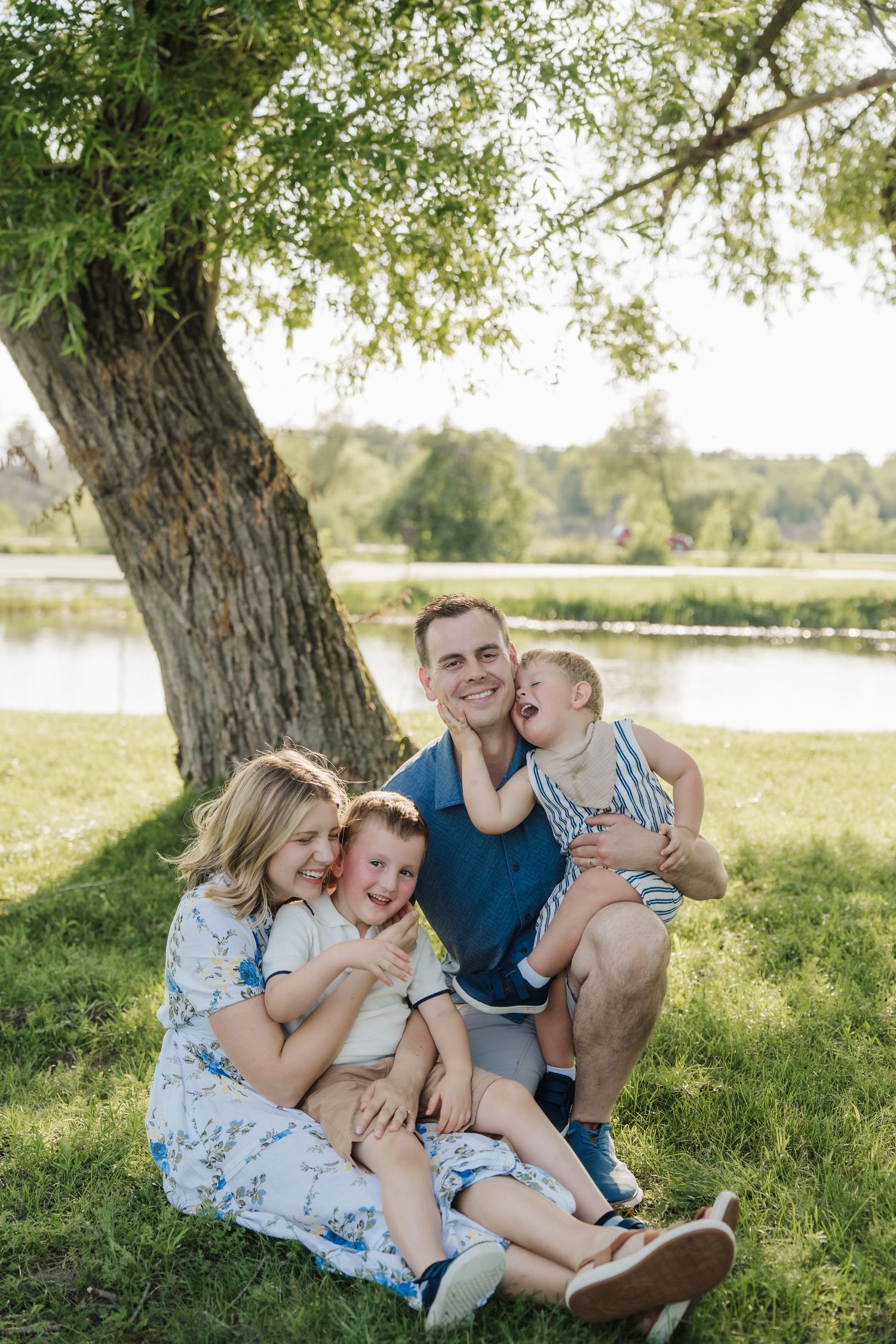 Happy family of four sitting under a large tree by a lake on a sunny day in a green park.