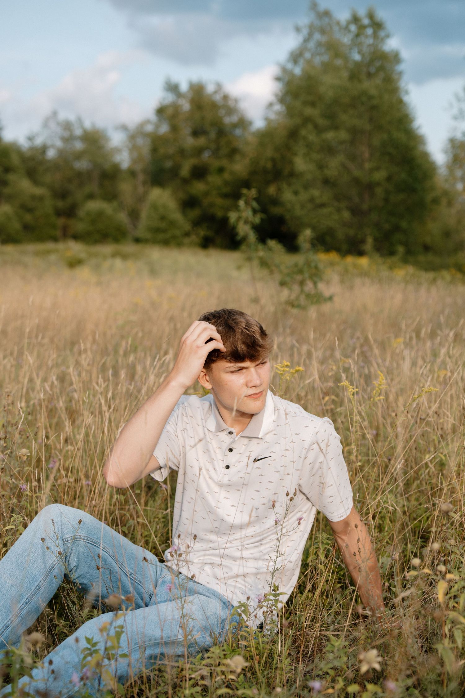 Young man in white Nike polo and jeans sitting in tall grass field with trees in background.