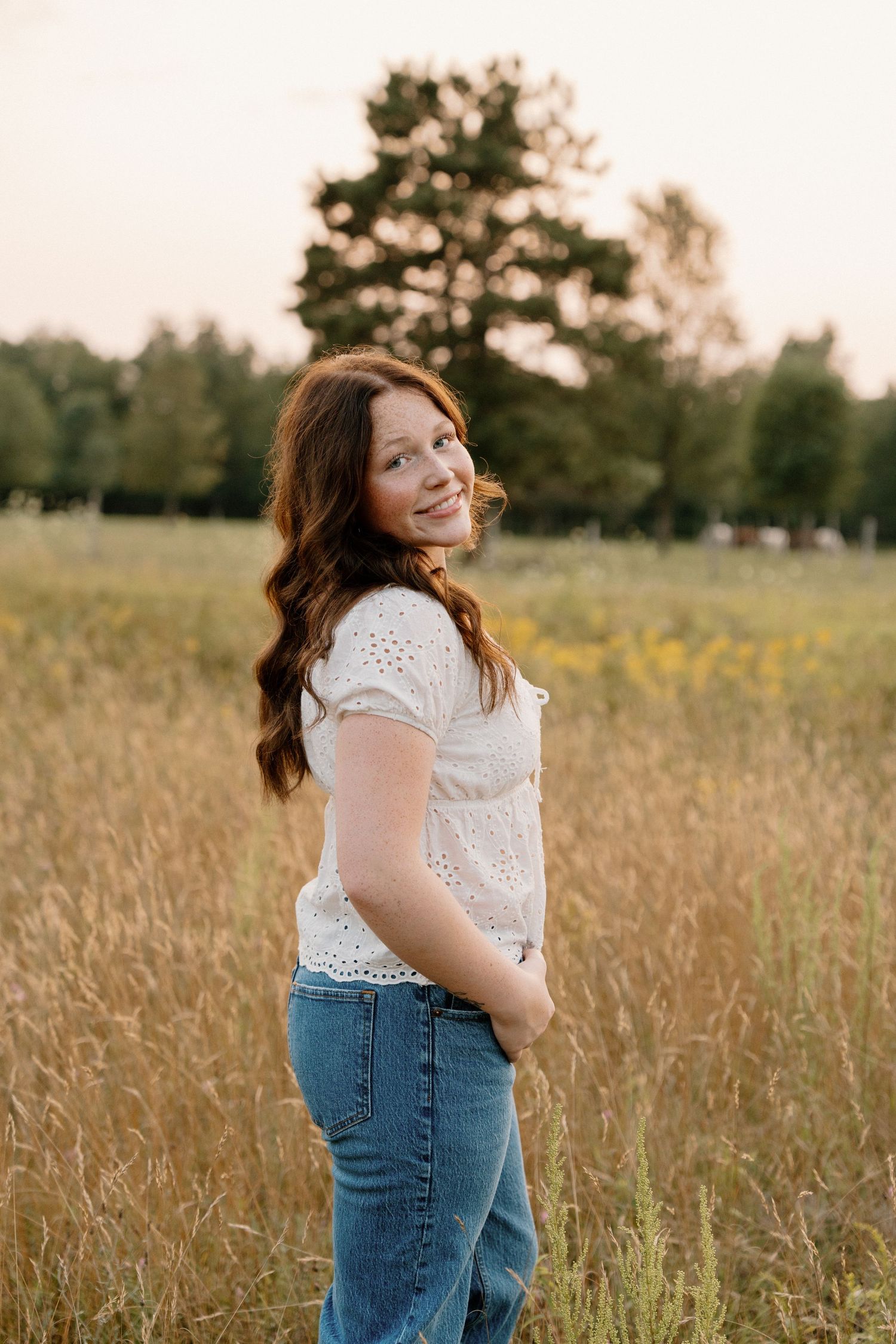 Young woman with red hair in white top and jeans smiling in golden field at sunset.