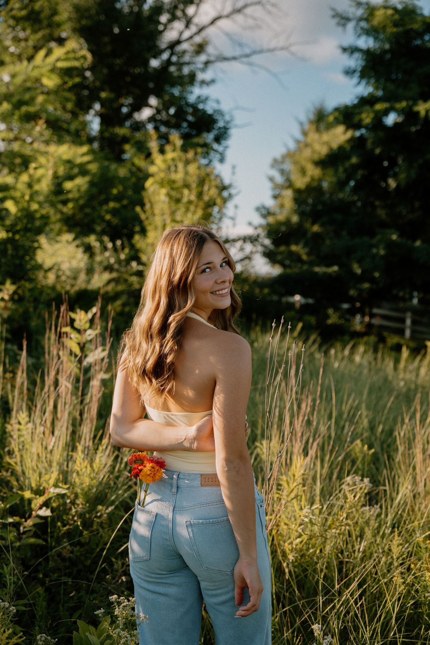 Young woman in crop top and jeans smiling over shoulder in sunny meadow with wildflowers and trees.
