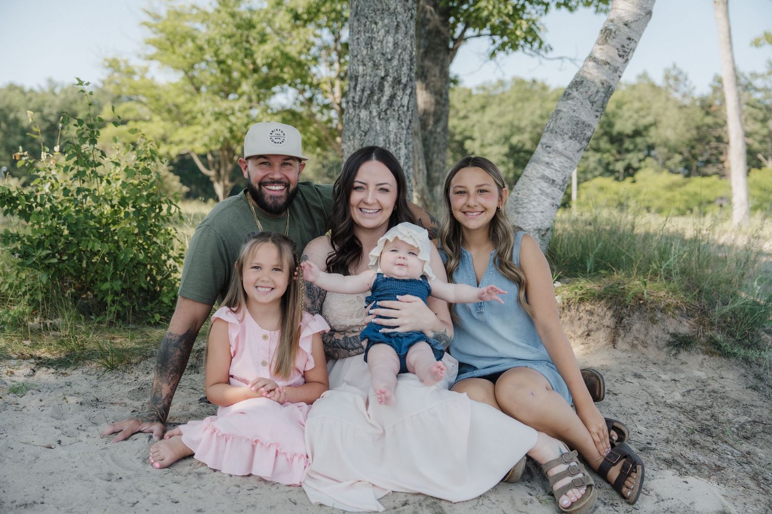 Happy family of five posing outdoors on sandy ground near trees, including two girls and a baby, smiling for a portrait.