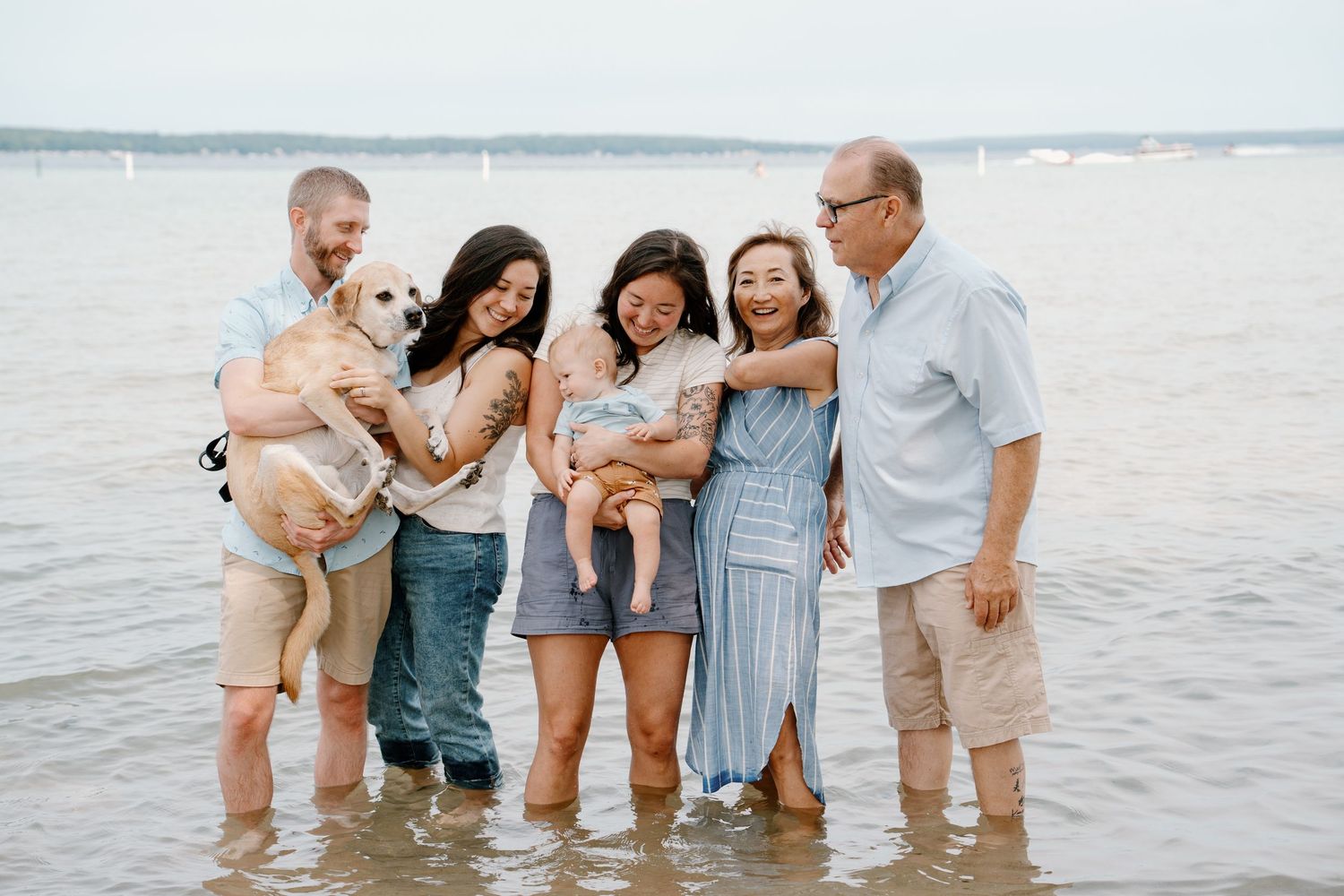 Family of five with a dog standing in shallow beach water, smiling and laughing together on a cloudy day.