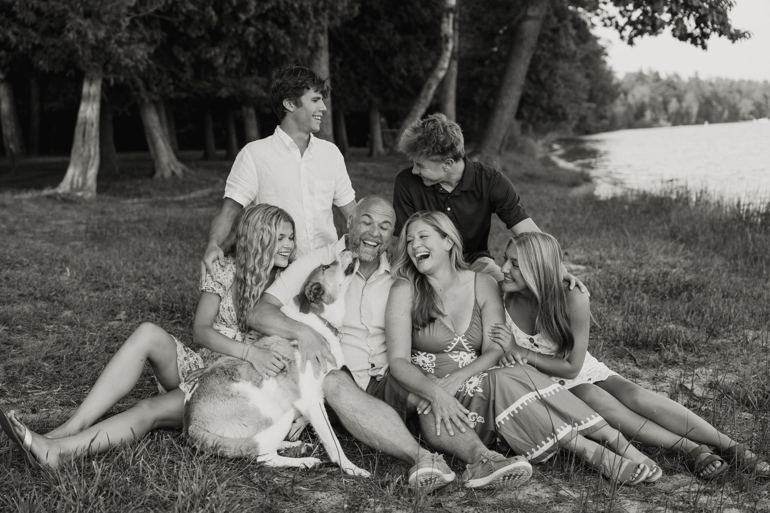 Black and white family photo of parents, children, and dog laughing together on grass near a lake.