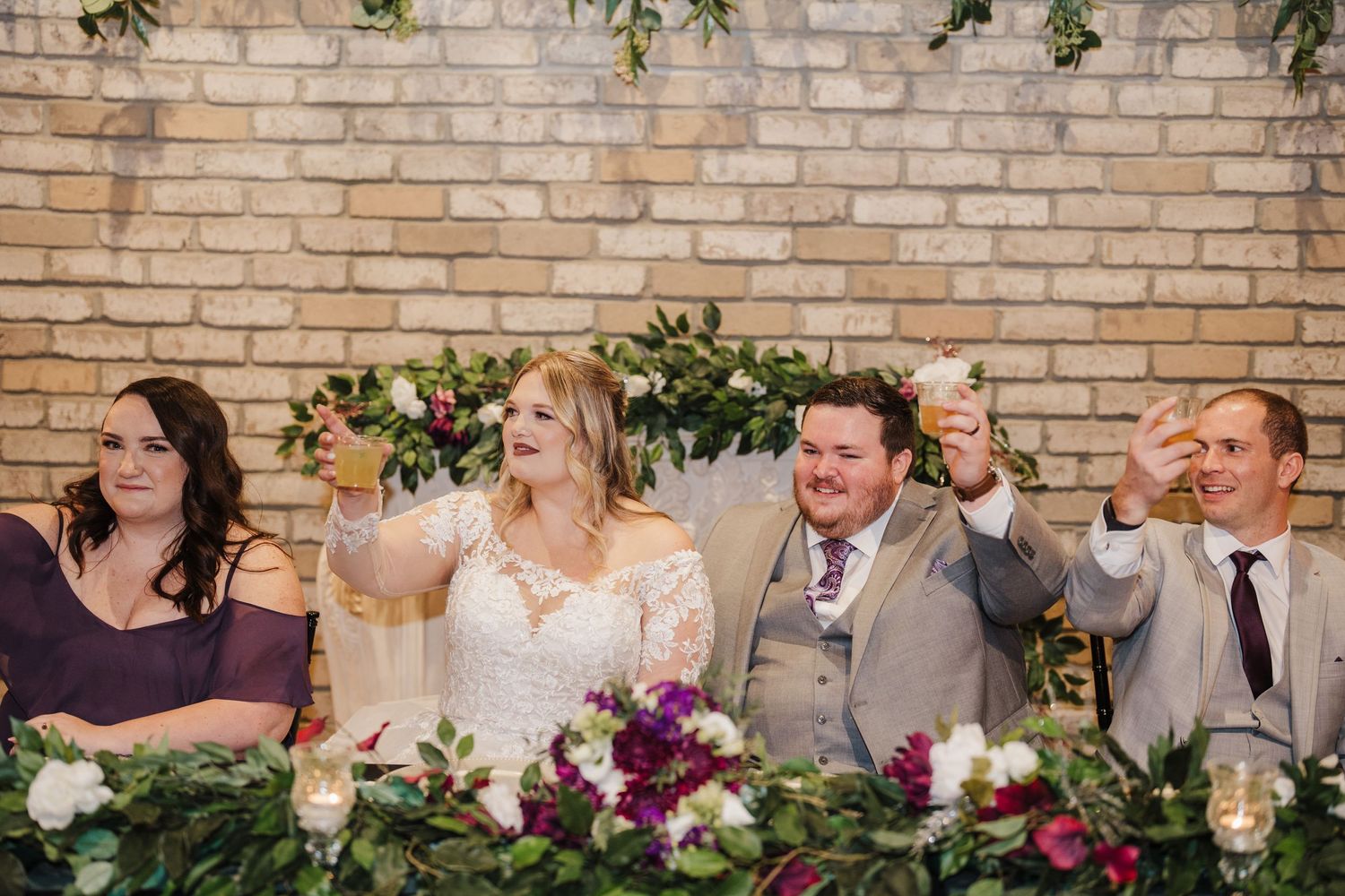 Wedding party celebrates at head table decorated with purple floral arrangements.