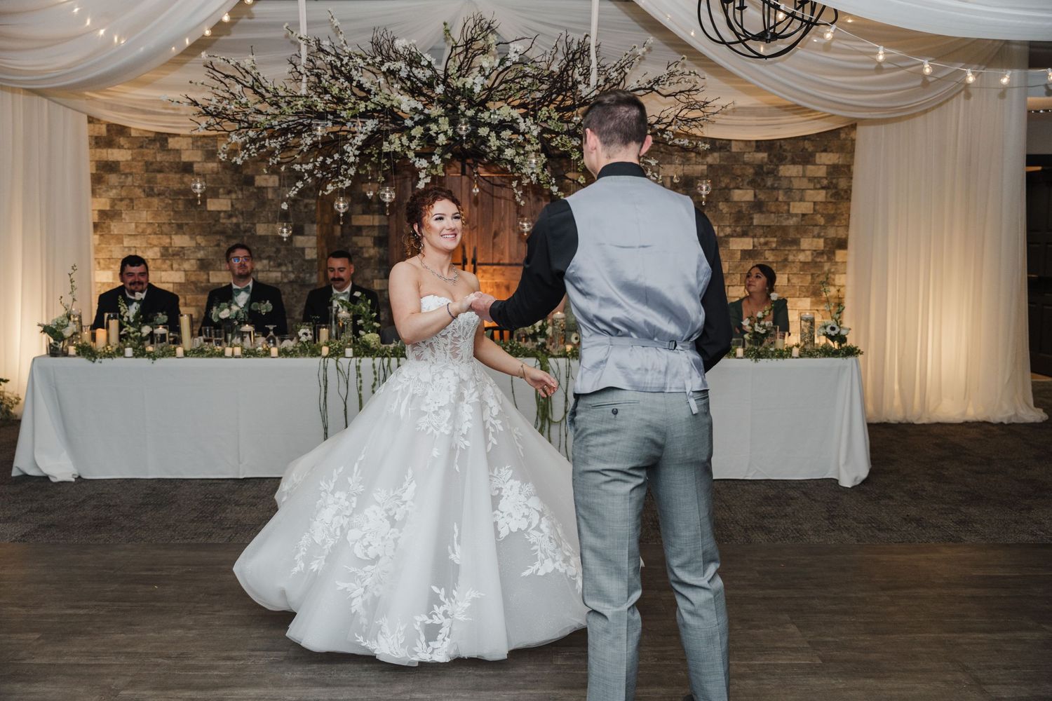 Couple shares first dance in reception hall decorated with floral arrangements and draped fabric.
