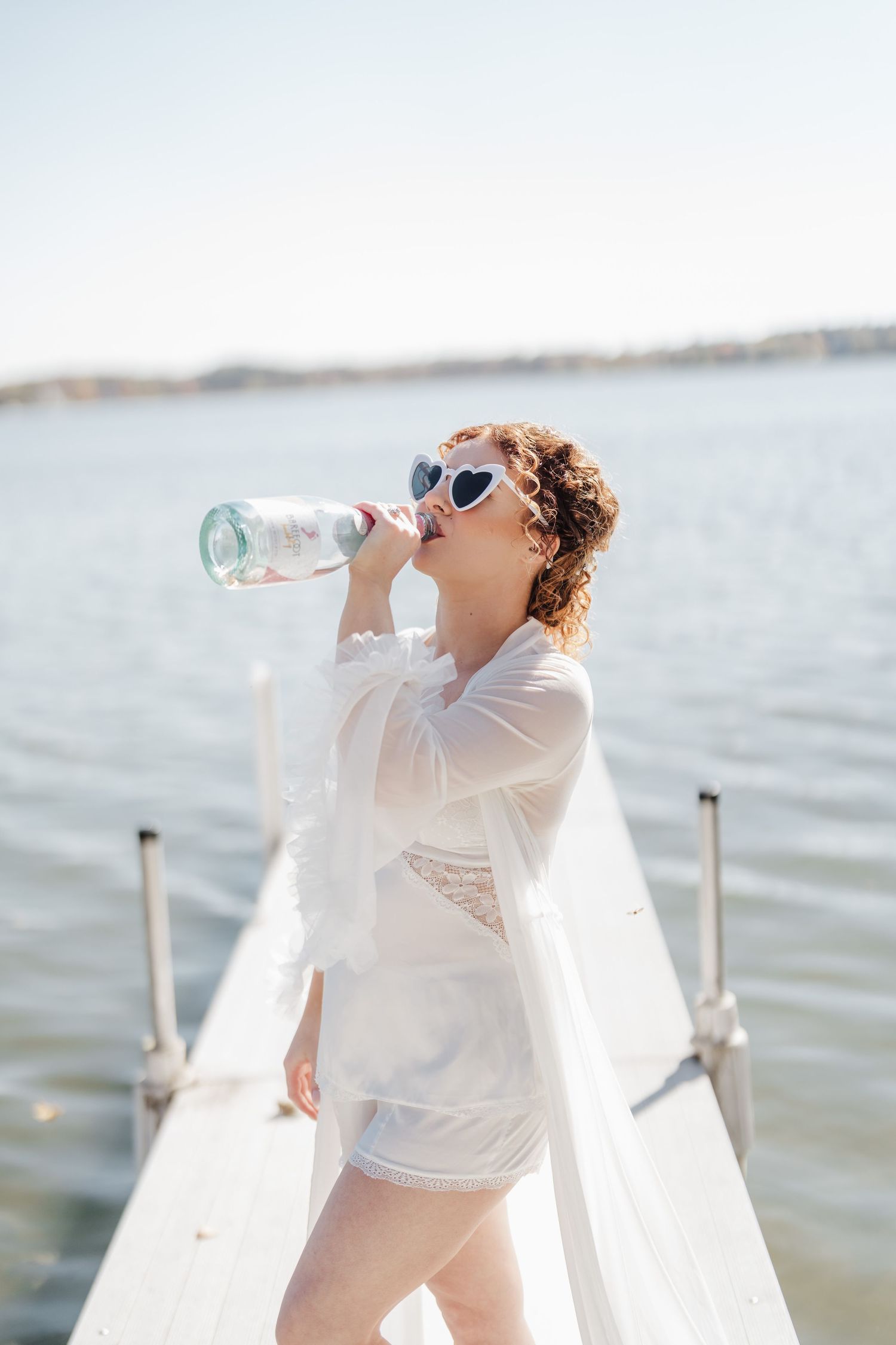 Woman in white coverup drinking from water bottle on dock by the ocean.