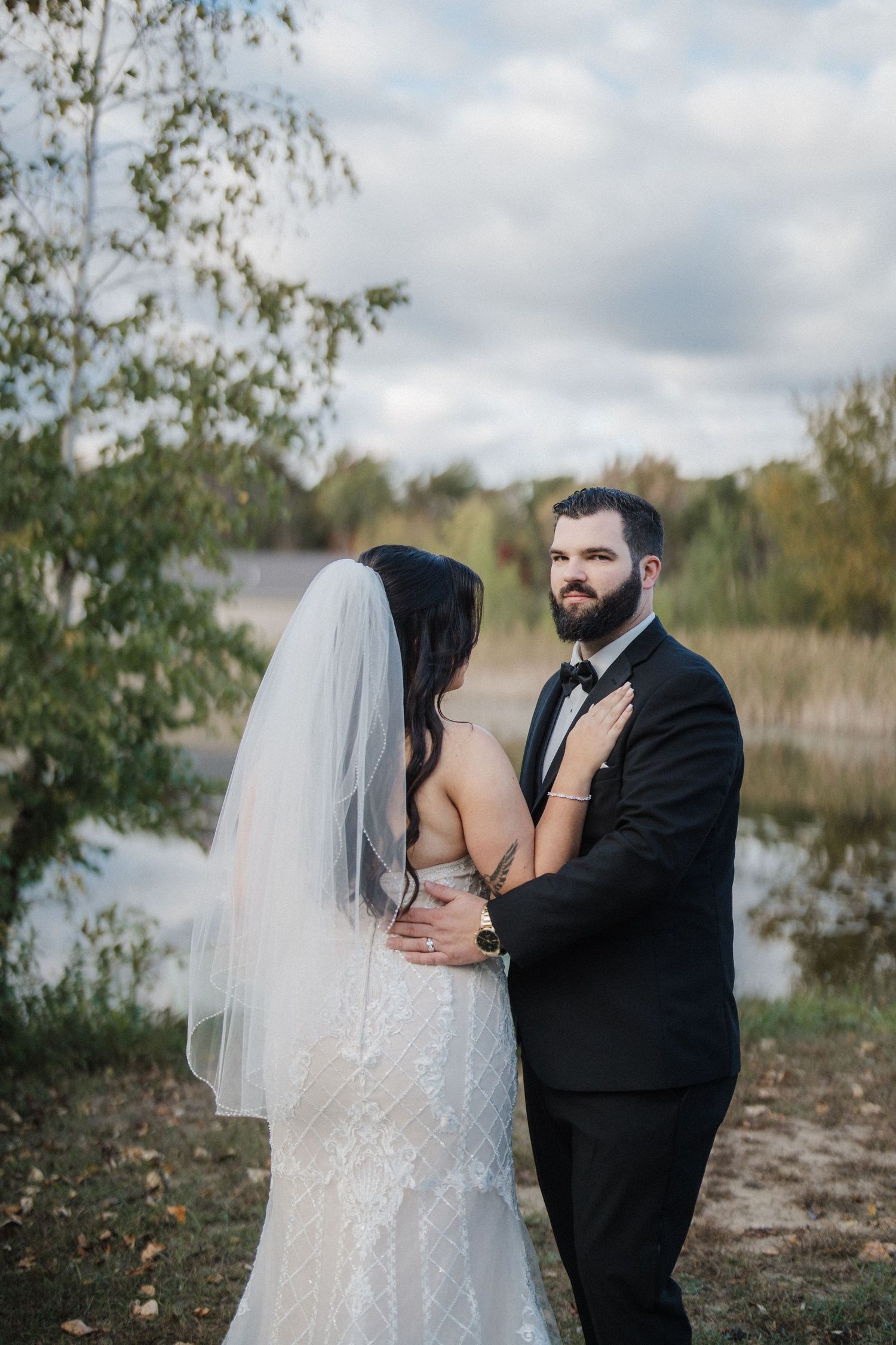 A couple shares an intimate moment outdoors by a lake during their wedding photoshoot on an autumn day.