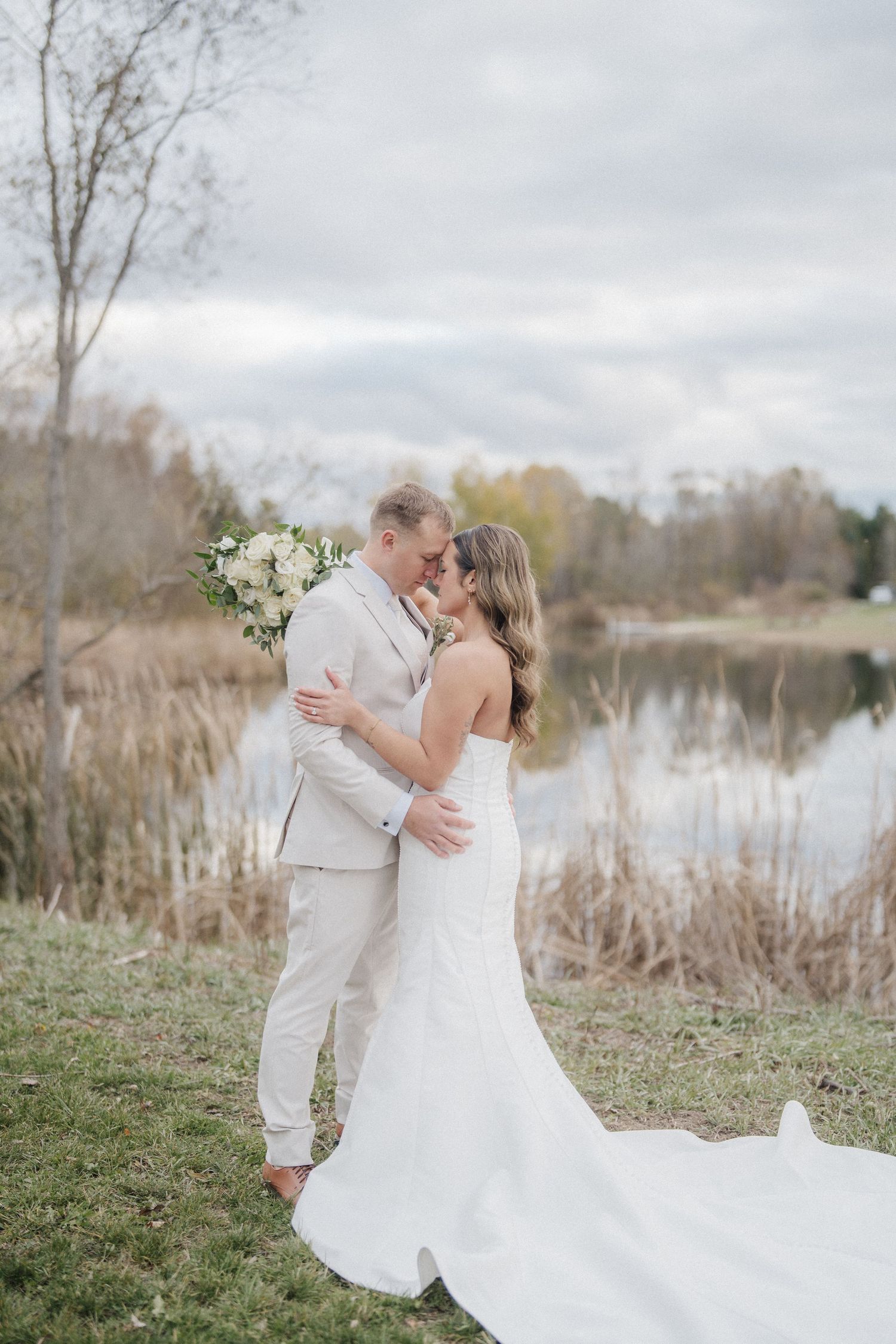 Newlyweds share intimate moments by a lake with cattails and autumn trees in the background.