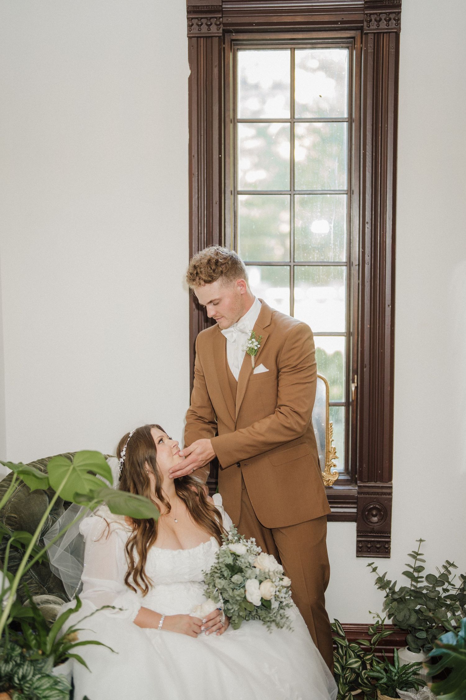Romantic wedding couple shares intimate moments in elegant indoor setting with natural light and greenery.