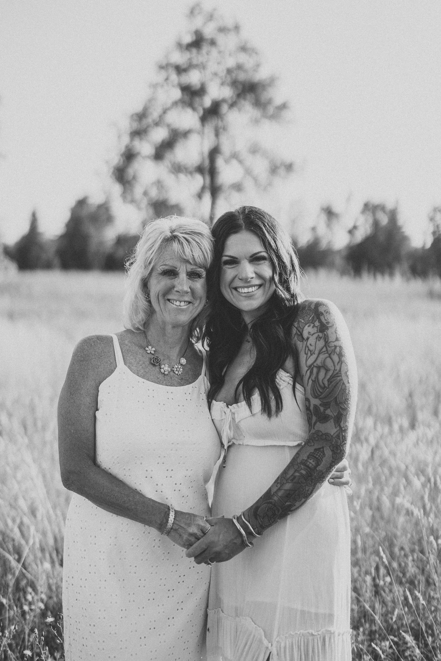 Two women in white dresses embrace while standing in a sunny field with trees in the background.
