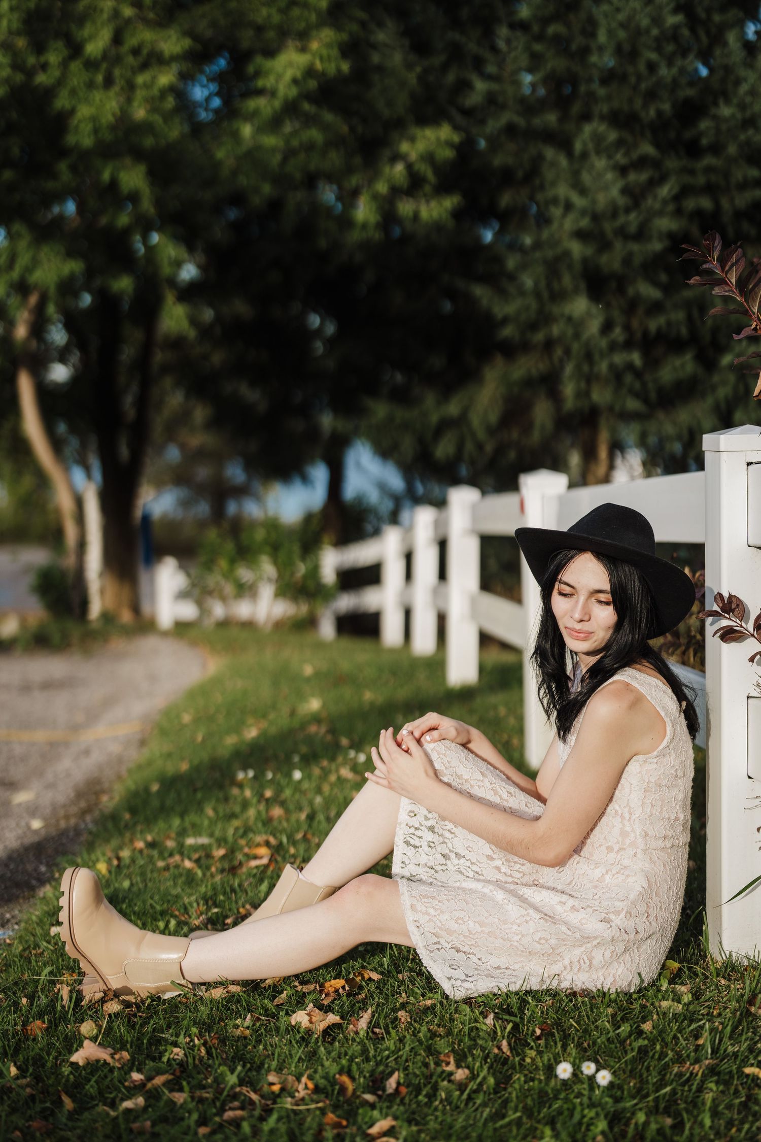 A person in a white lace dress and black hat sits against a white fence on grass with autumn leaves scattered around.