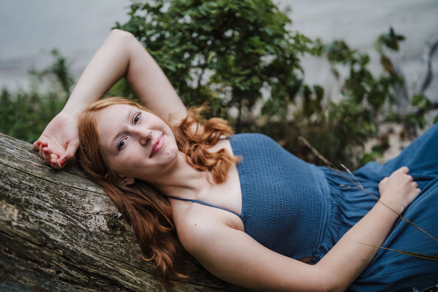 Person with red hair wearing a blue dress reclines on a fallen tree trunk in a natural outdoor setting.