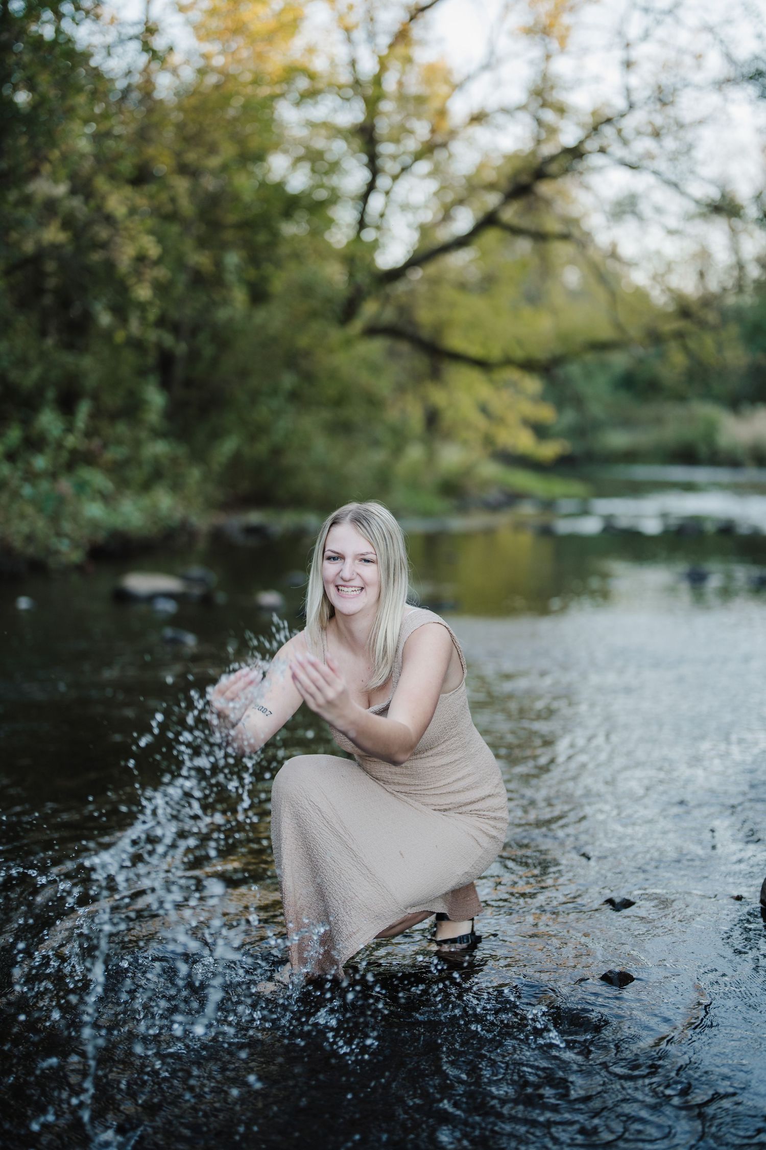 A person in a beige dress splashes water while sitting on rocks by a scenic river surrounded by trees.