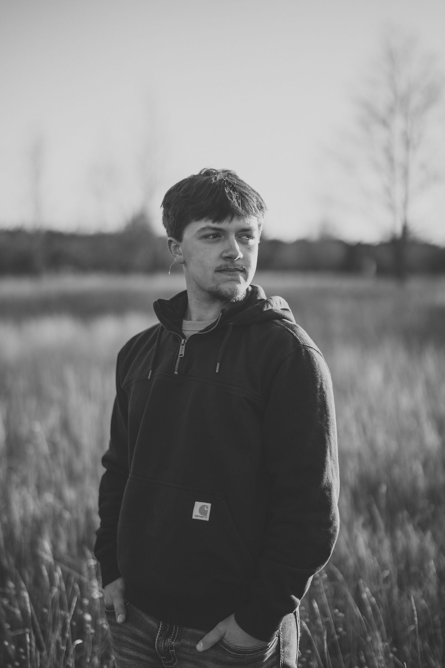 Black and white portrait of person in Carhartt hoodie standing in tall grass field at dusk.