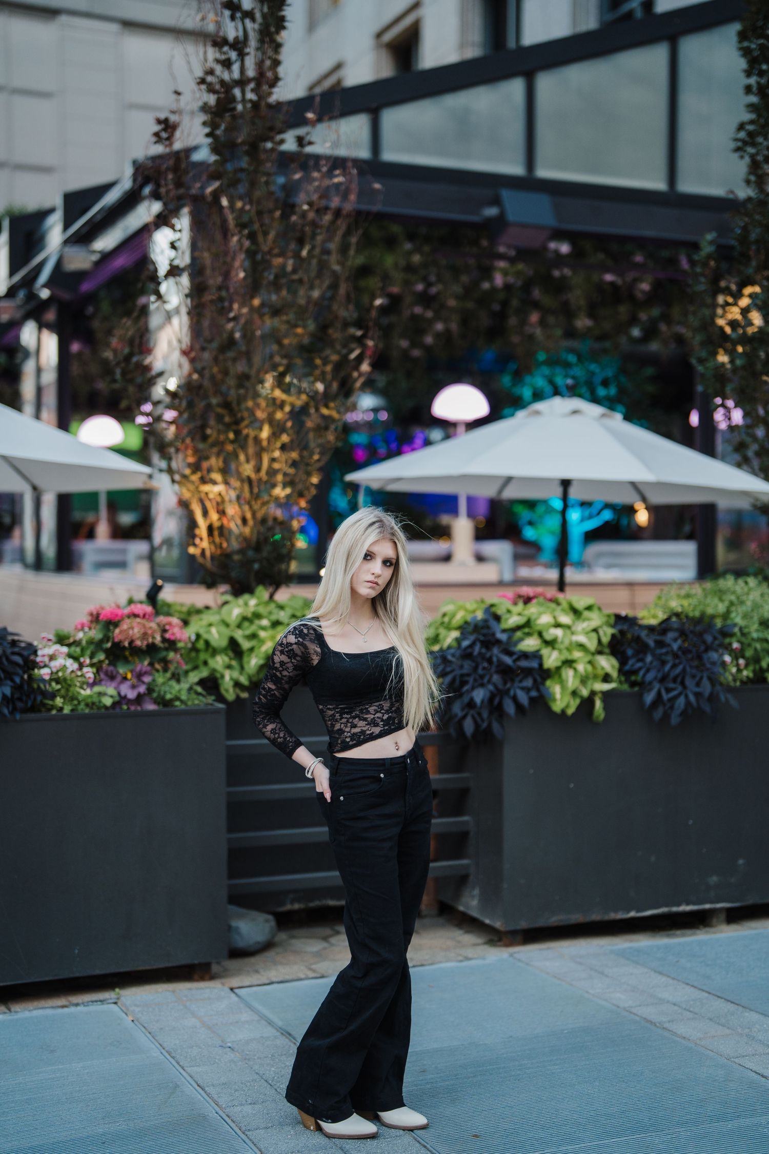 A figure in an elegant black dress poses gracefully against a backdrop of urban planters and umbrellas.