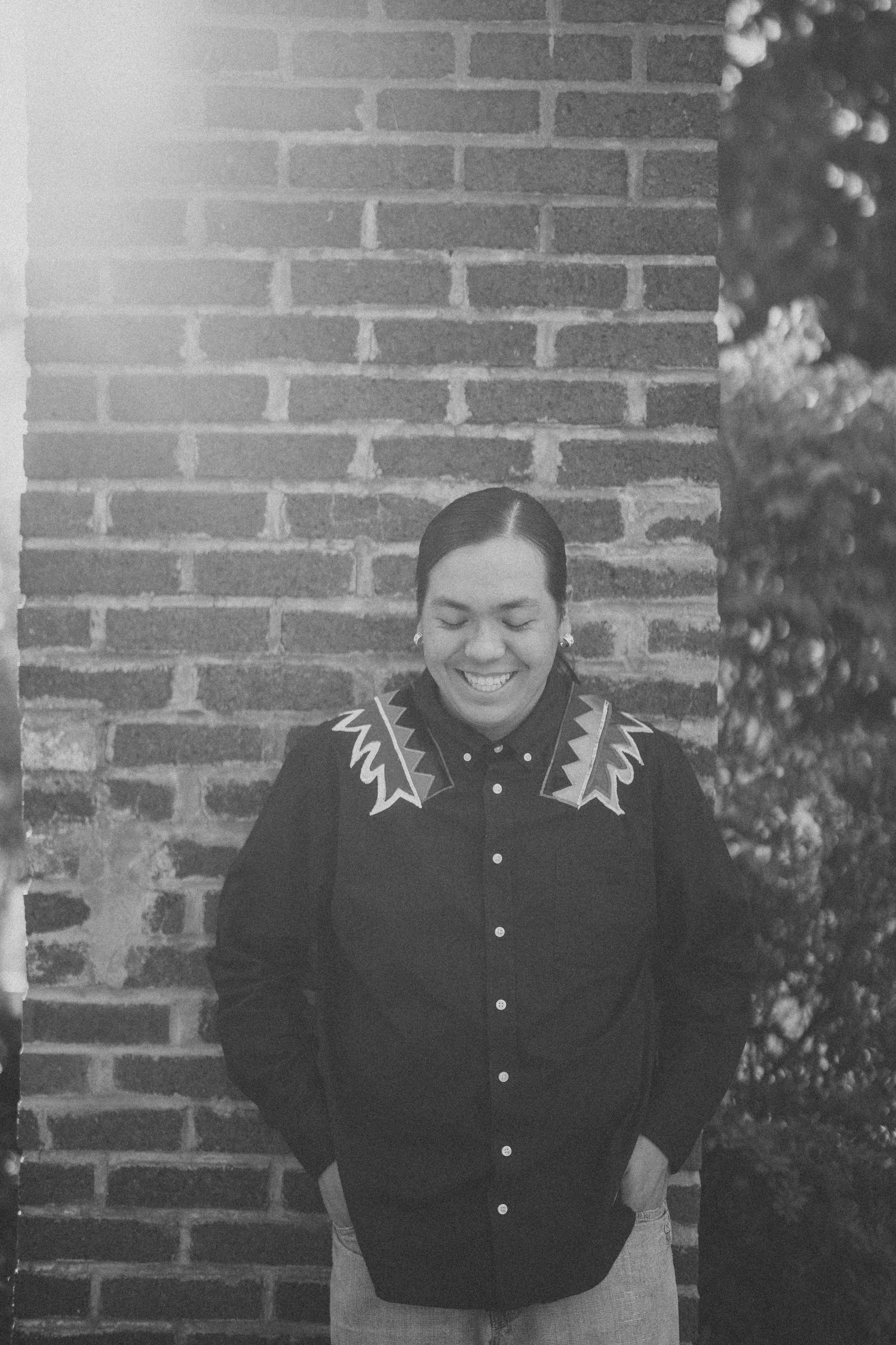 A person wearing a patterned cardigan smiles joyfully while standing against a brick wall in black and white.