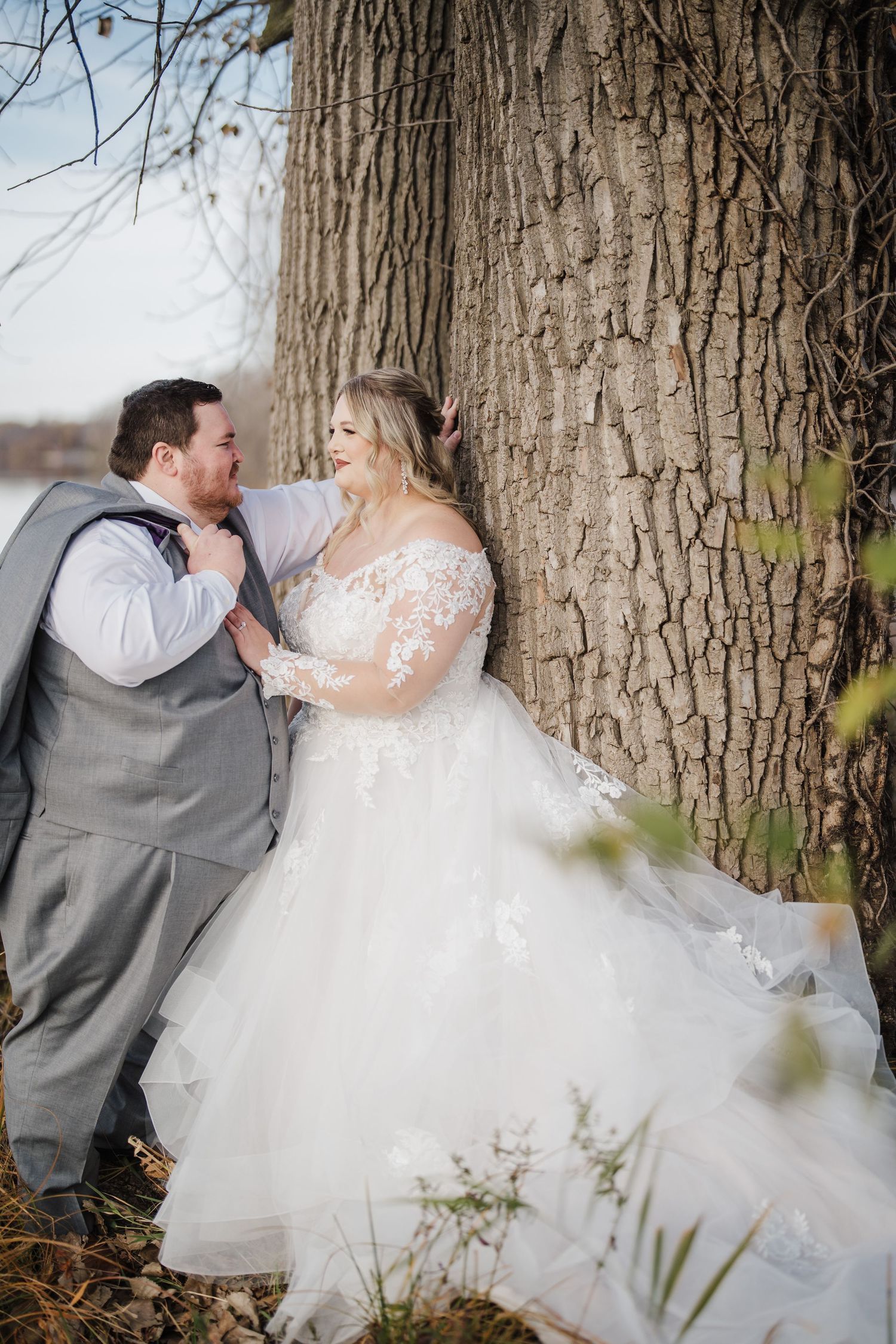 Newlyweds share an intimate moment against a large tree trunk in their wedding attire.