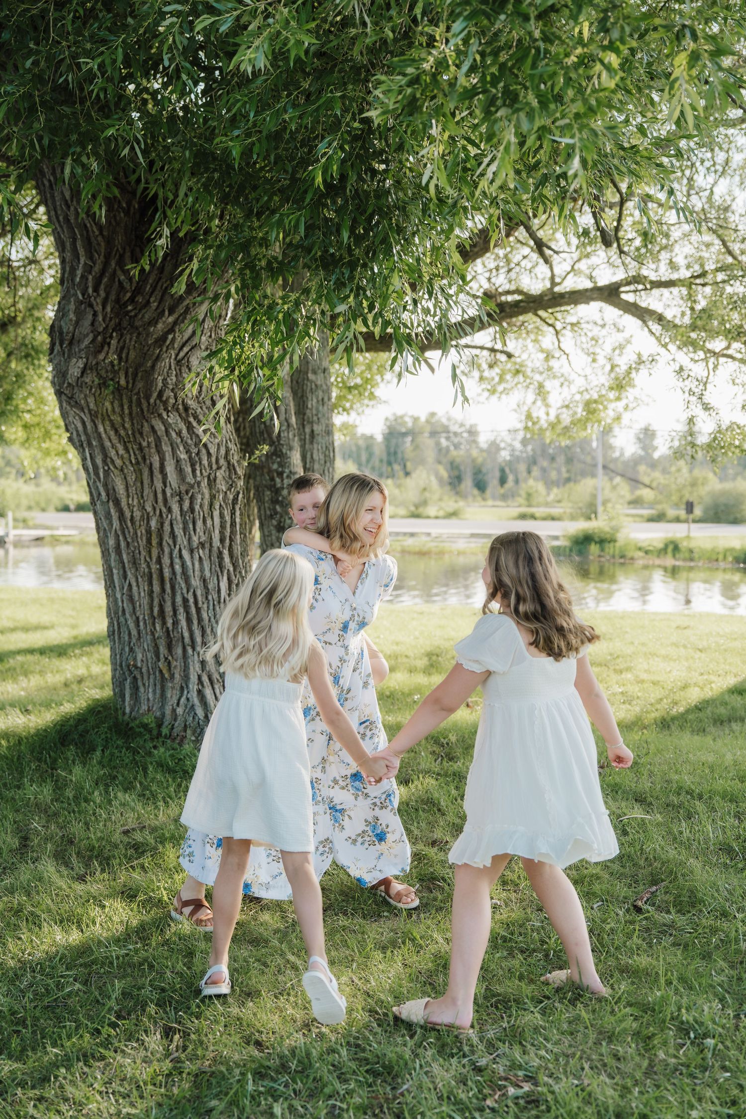 A mother in a floral dress holds hands with two girls in white dresses, dancing under a large tree by a pond.