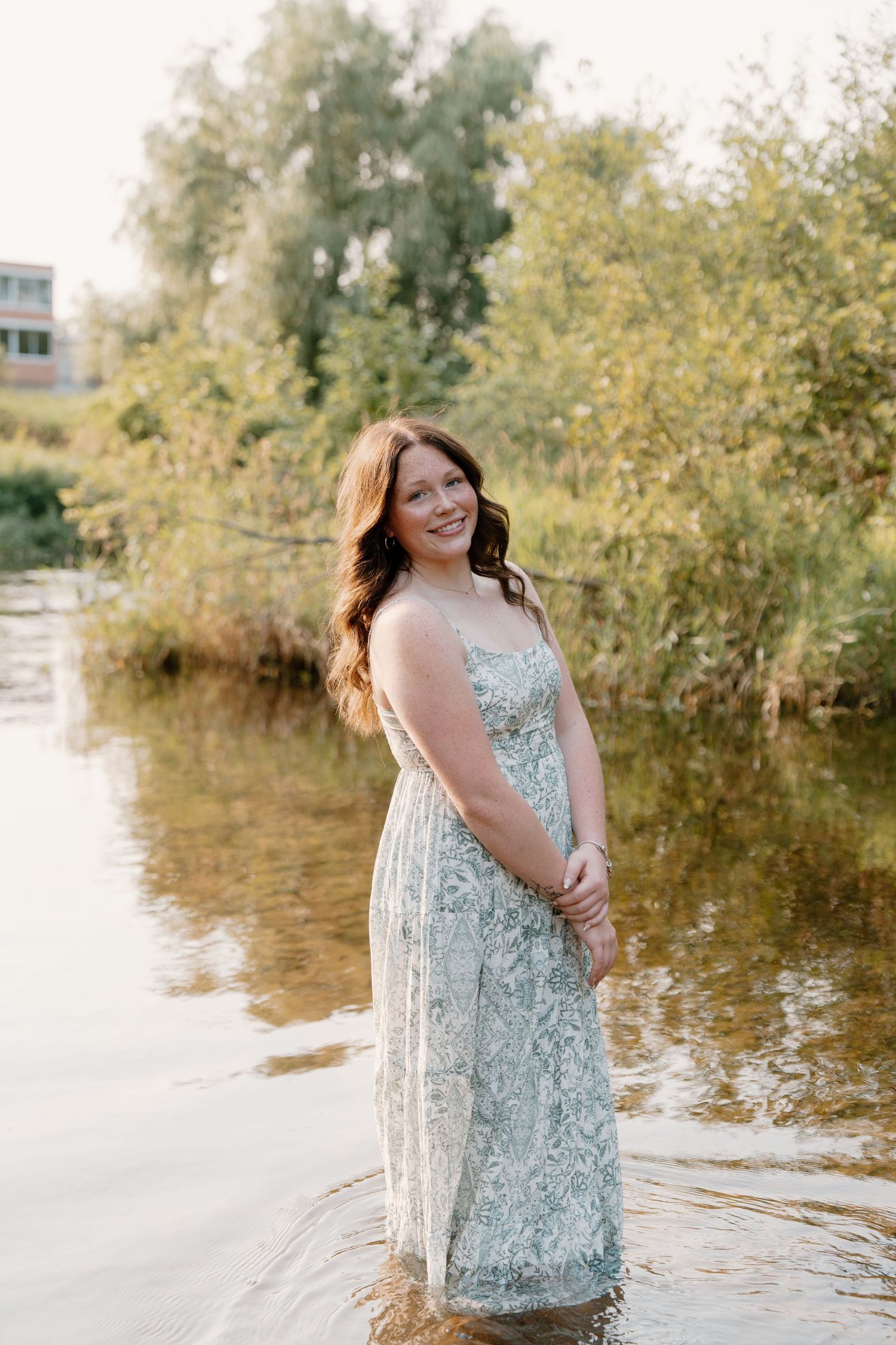 Young woman in floral mint dress standing in shallow river water, smiling, with lush green trees in background.