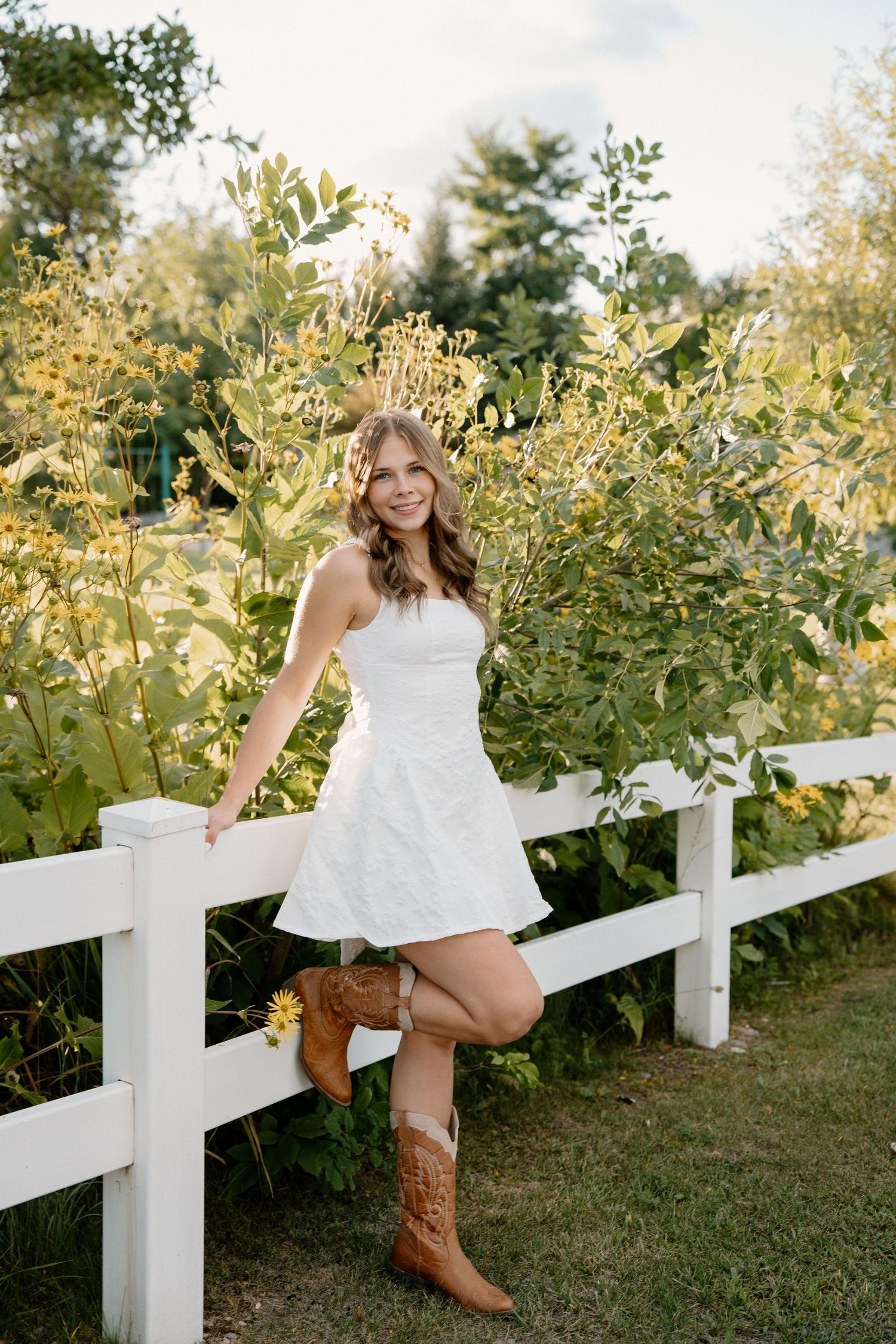 Young woman in white strapless dress and brown cowboy boots leaning on white fence surrounded by lush greenery.