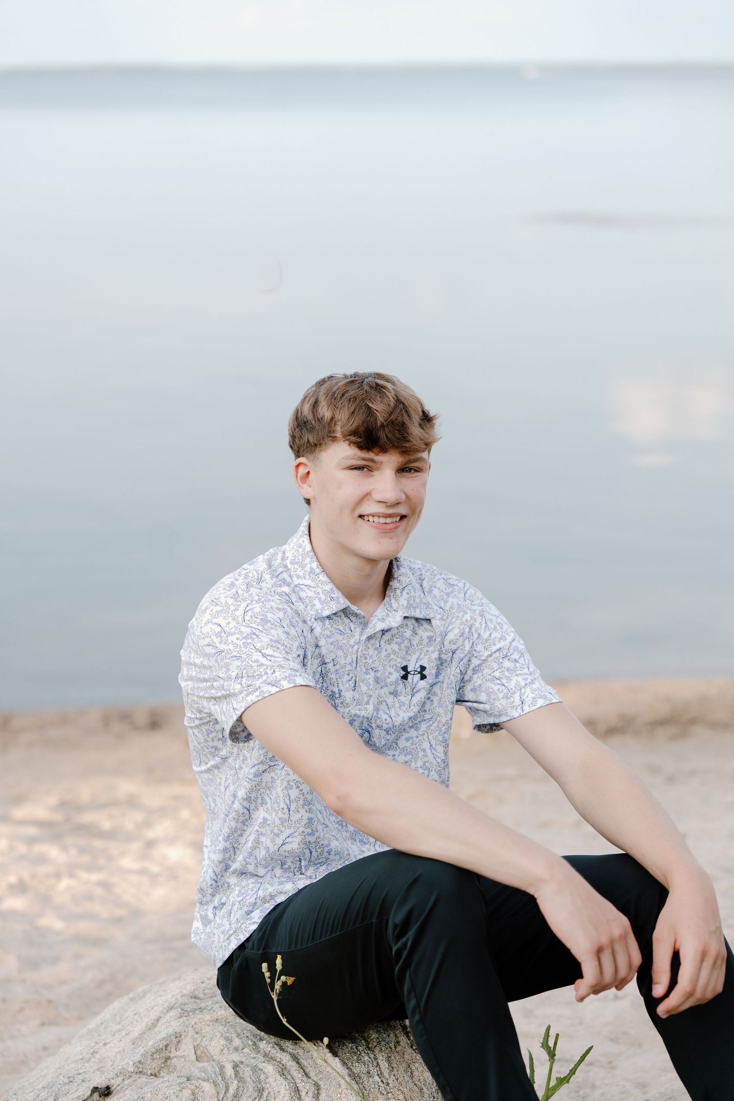 Smiling teen boy in white Under Armour polo sits on rock at sandy beach with calm water behind him.
