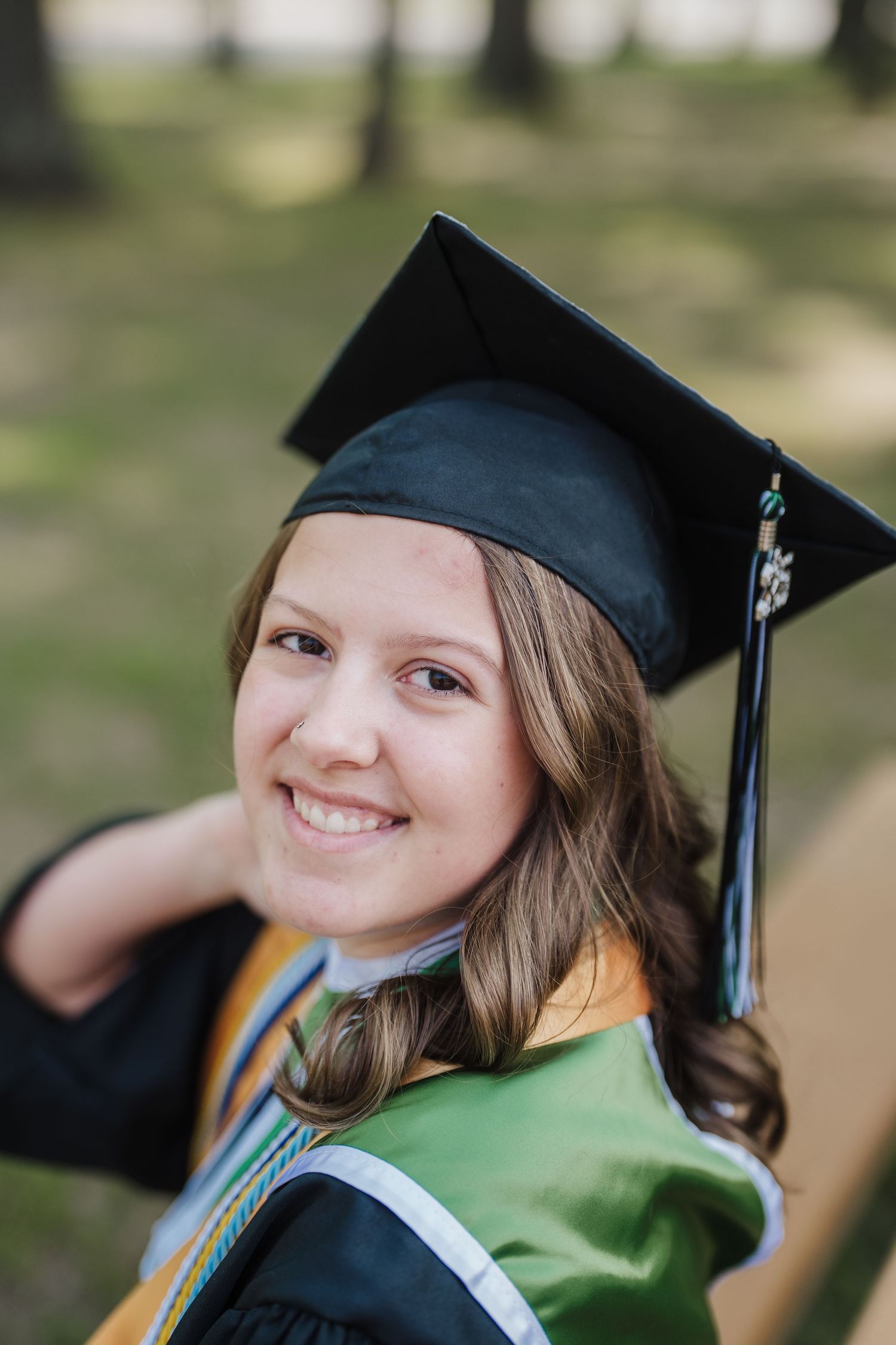 Graduate in black cap and gown with honors cords poses outdoors on graduation day.