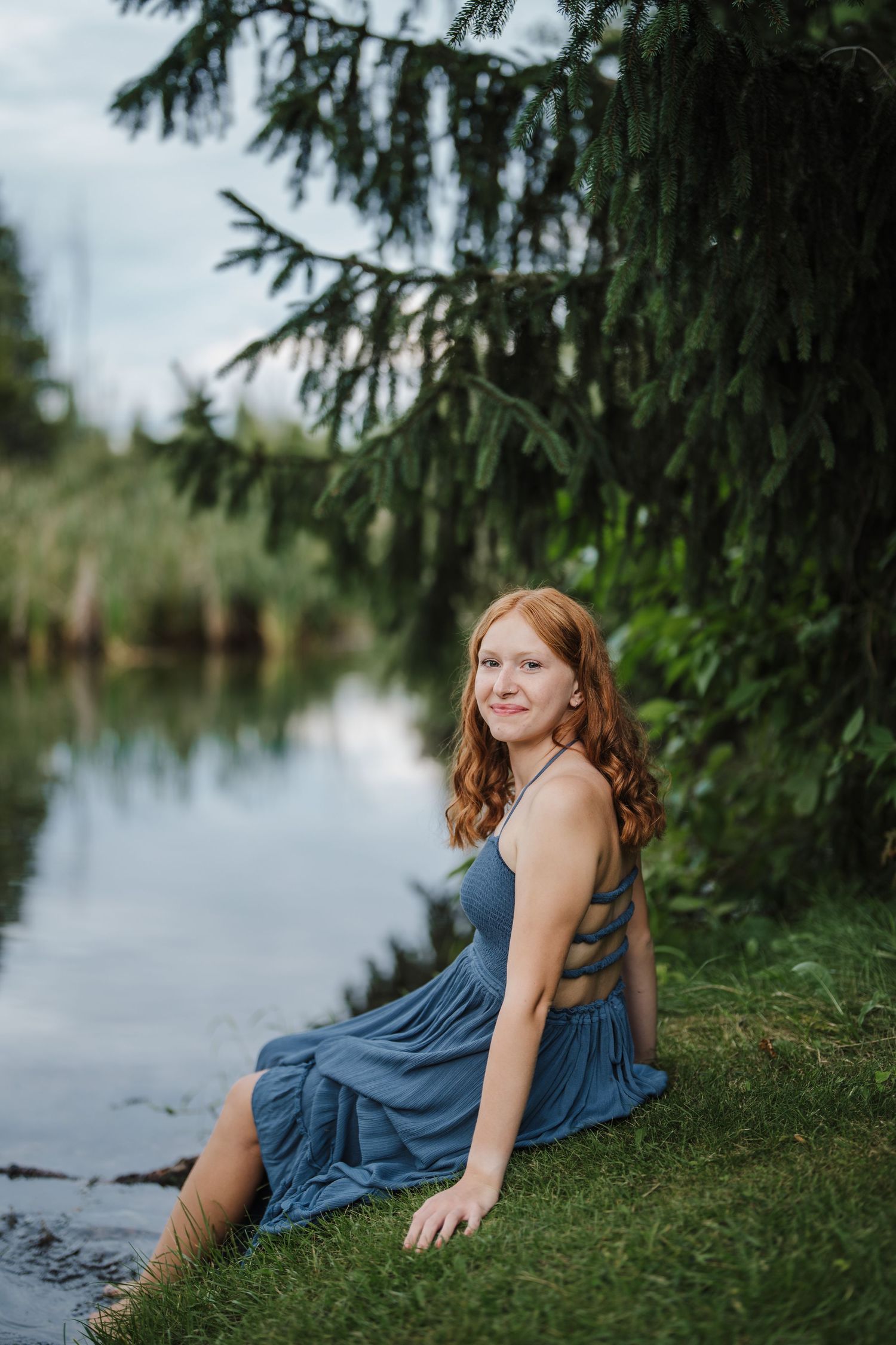 A person in a blue dress sits by a tranquil lake under evergreen branches on a peaceful summer day.