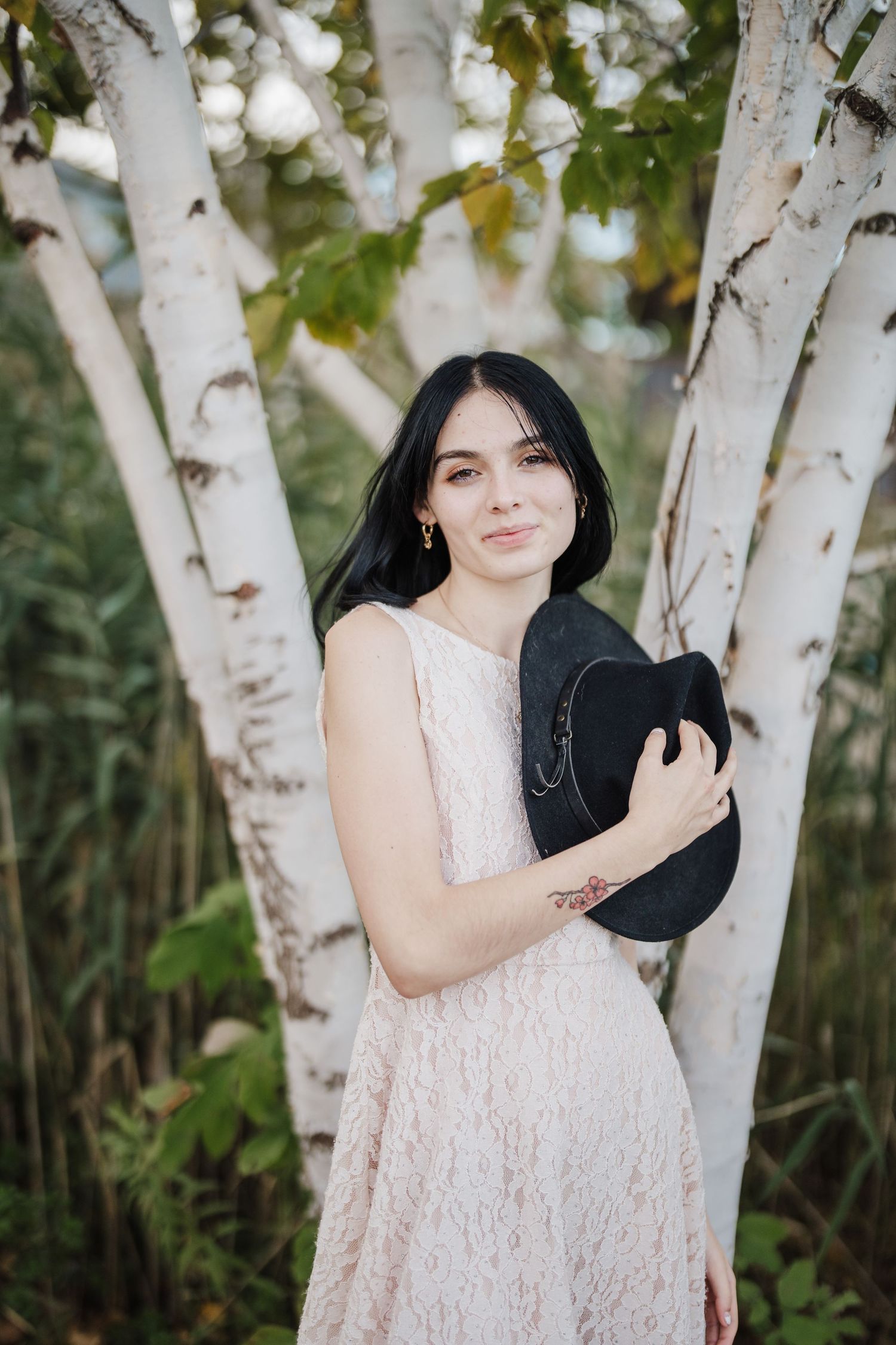 A person in a white lace dress poses gracefully against white birch trees in a natural outdoor setting.