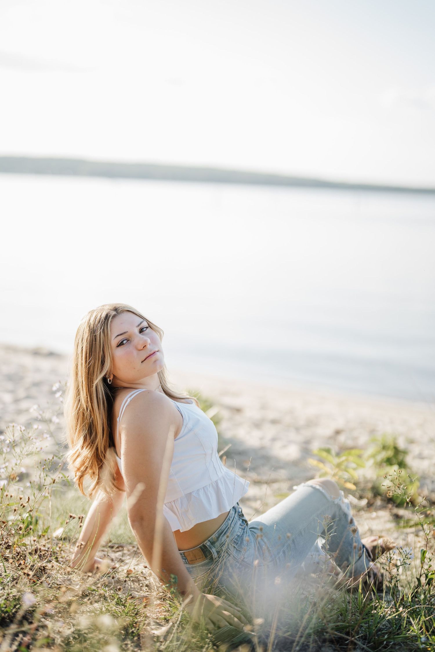 A person in a white top and jeans relaxes on a sunny beach shore with water and soft light in the background.