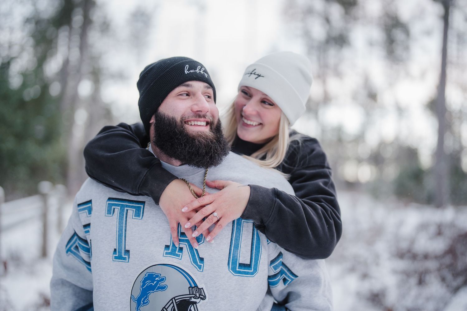 A couple embraces in a snowy winter setting wearing matching blue football jerseys and white beanies.