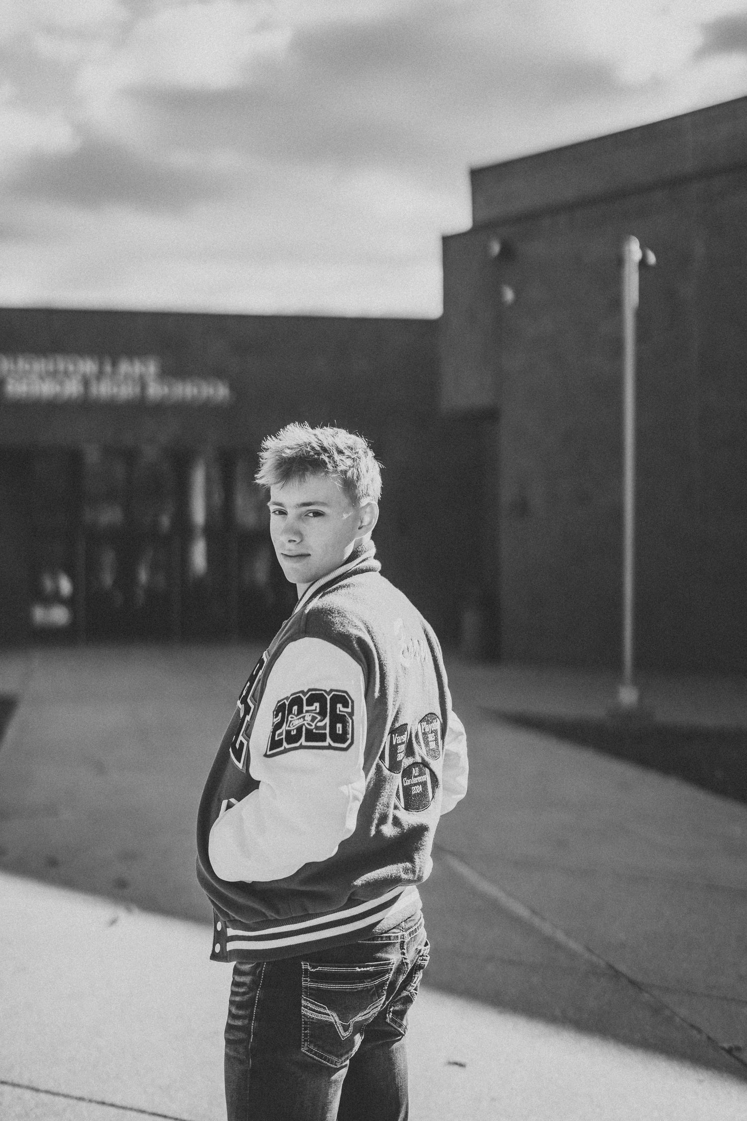 Black and white photo of student in varsity jacket standing outside school building.