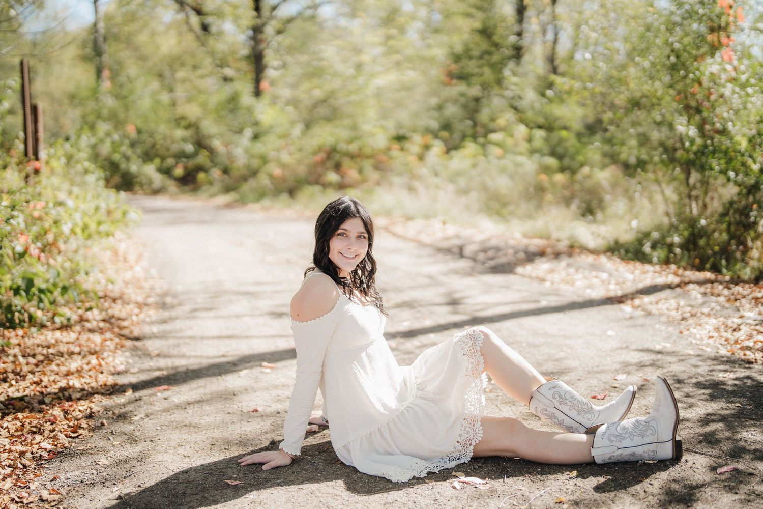 A person in a white dress reclines on a rustic dirt path surrounded by green foliage and dappled sunlight.