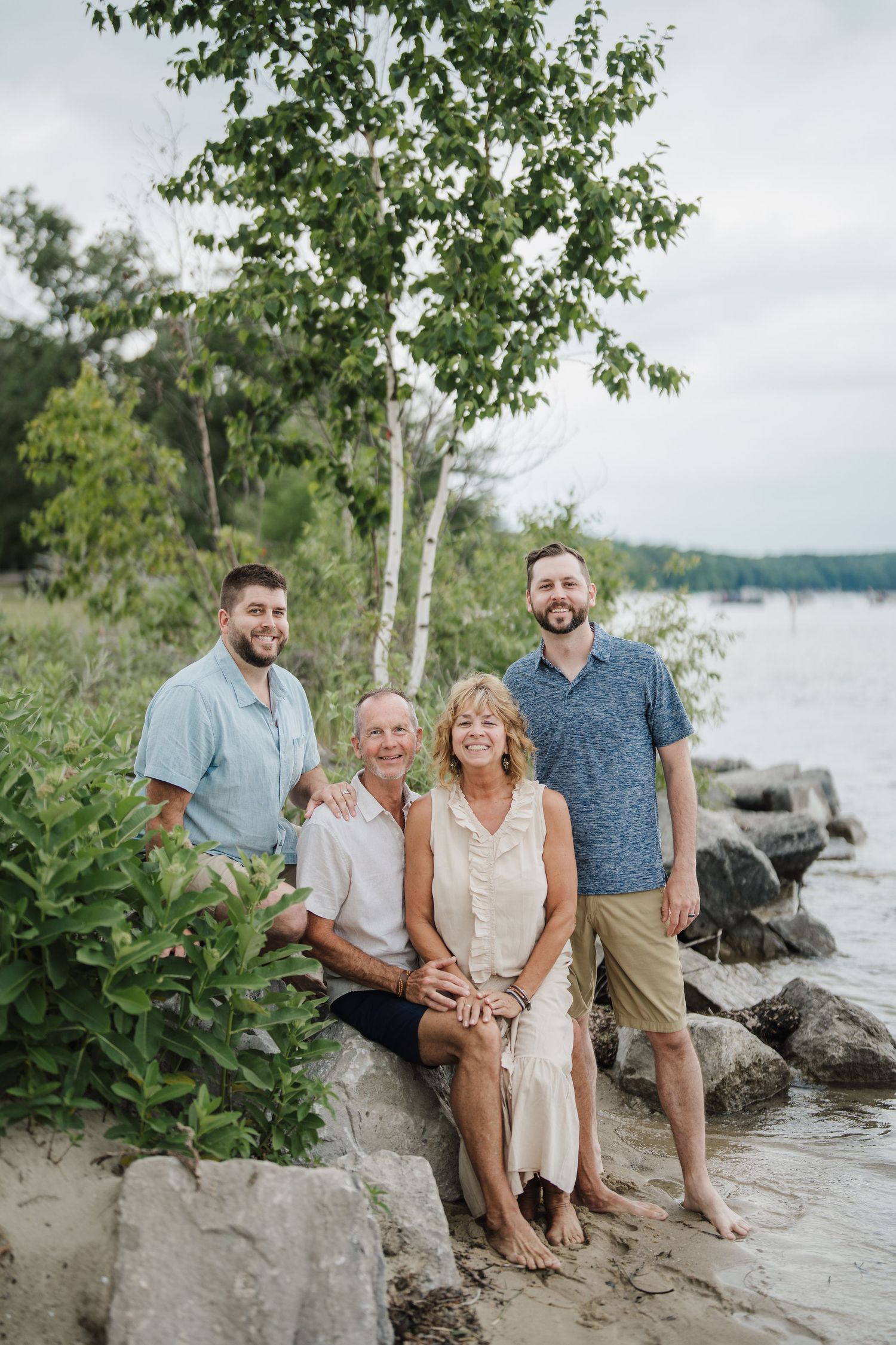 A family poses casually on rocky shoreline with trees and water in the background during an outdoor photo session.