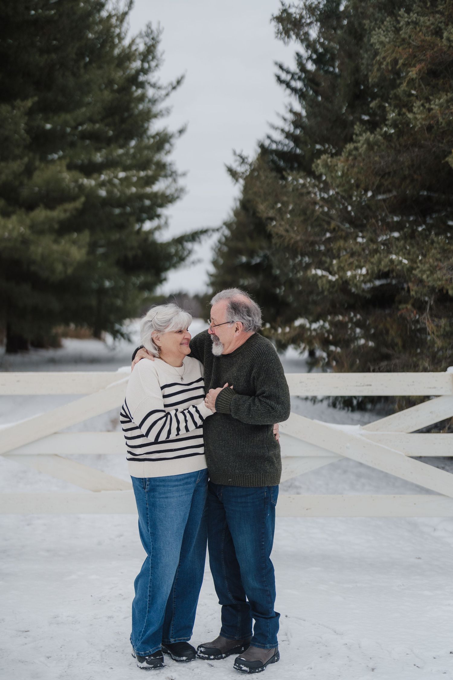 Elderly couple dancing together playfully in the snow with evergreen trees in the background.