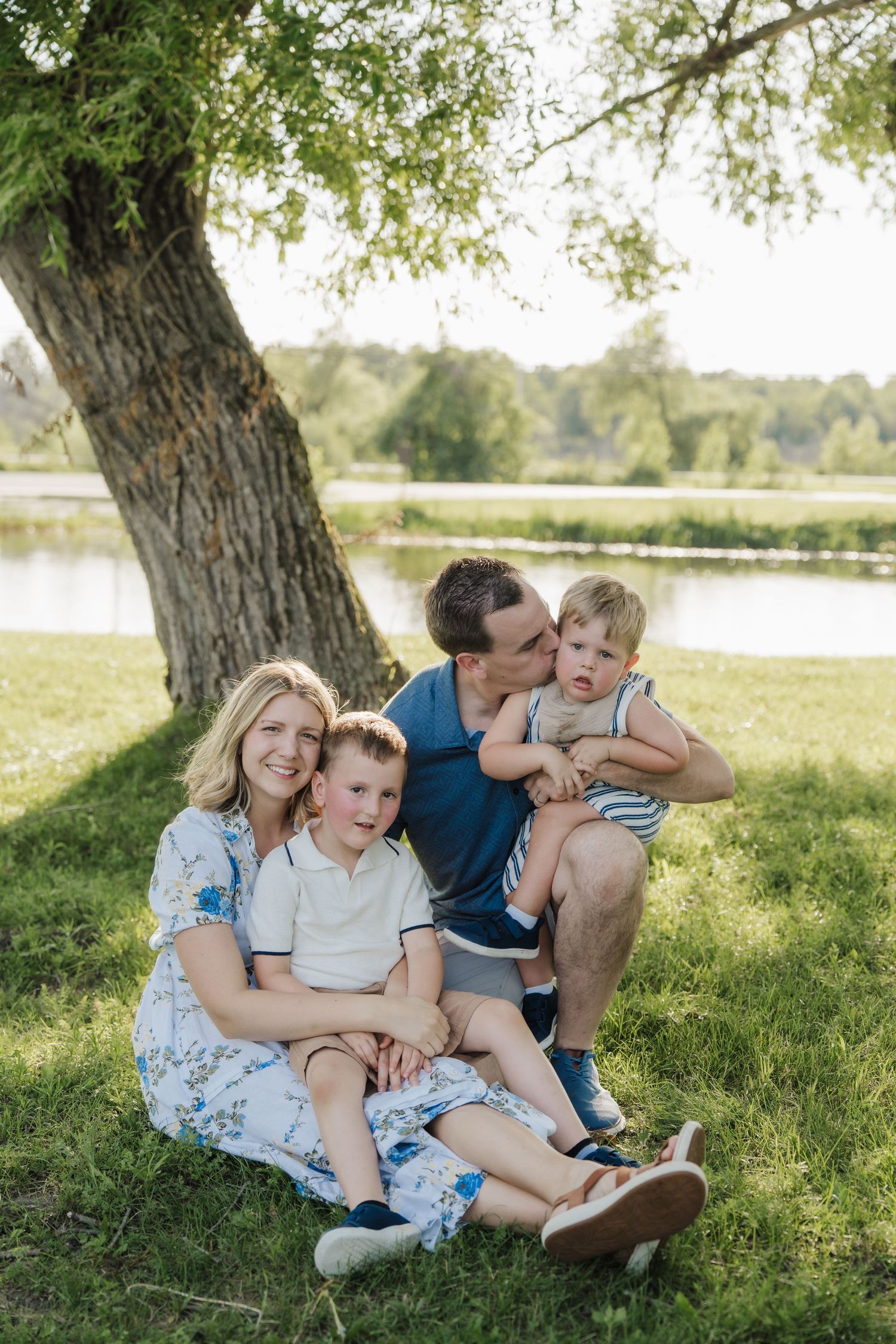 Happy family of four sitting on grass under a tree by a lake on a sunny day.