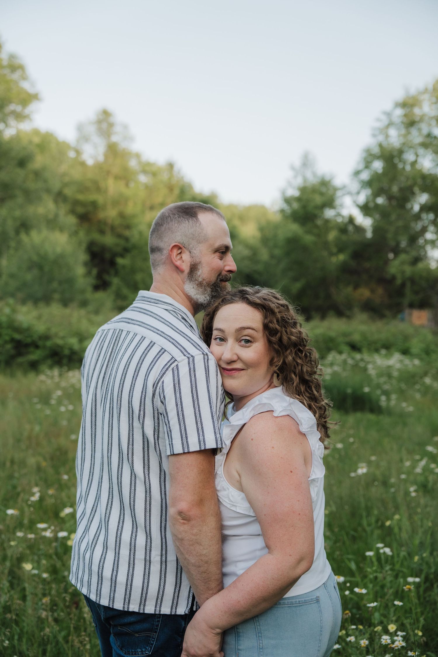 Couple embracing in a green meadow, woman smiling at camera, man in striped shirt, outdoor engagement photo.