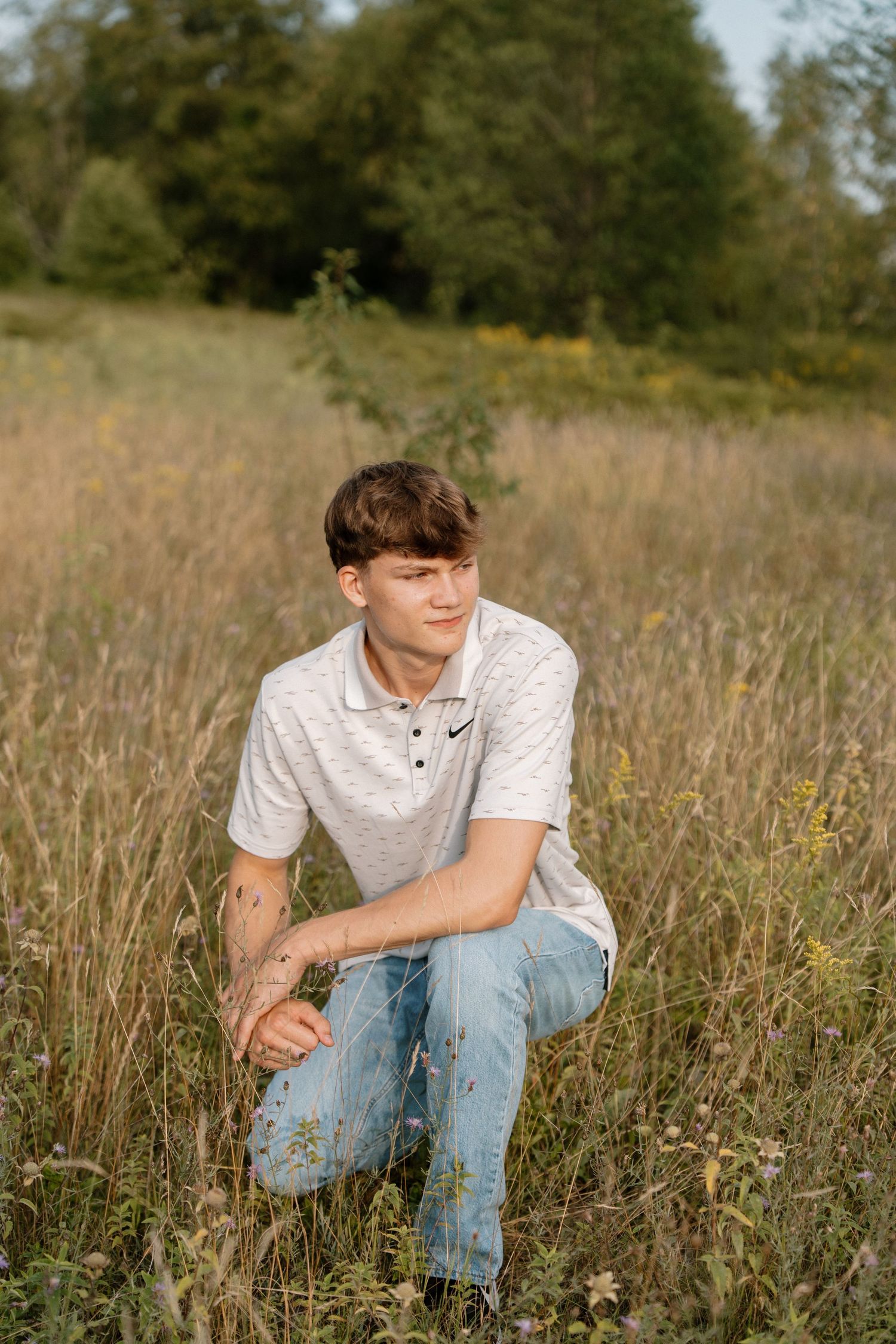 Young man in white Nike polo and jeans crouching in a golden meadow with wildflowers and trees behind.