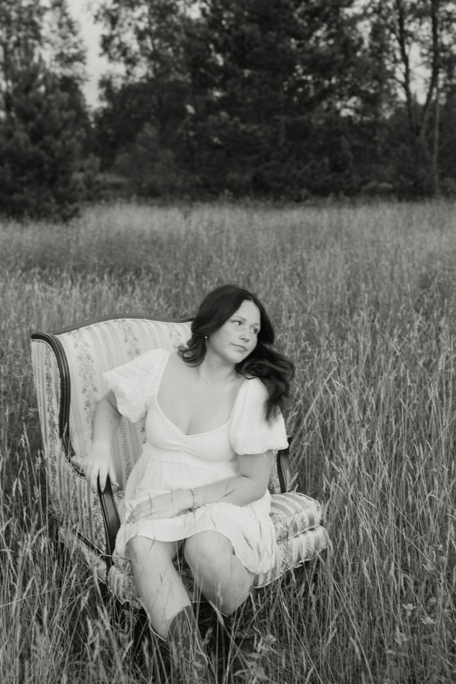 Black and white photo of a young woman in a white dress sitting in a vintage chair in a grassy field.