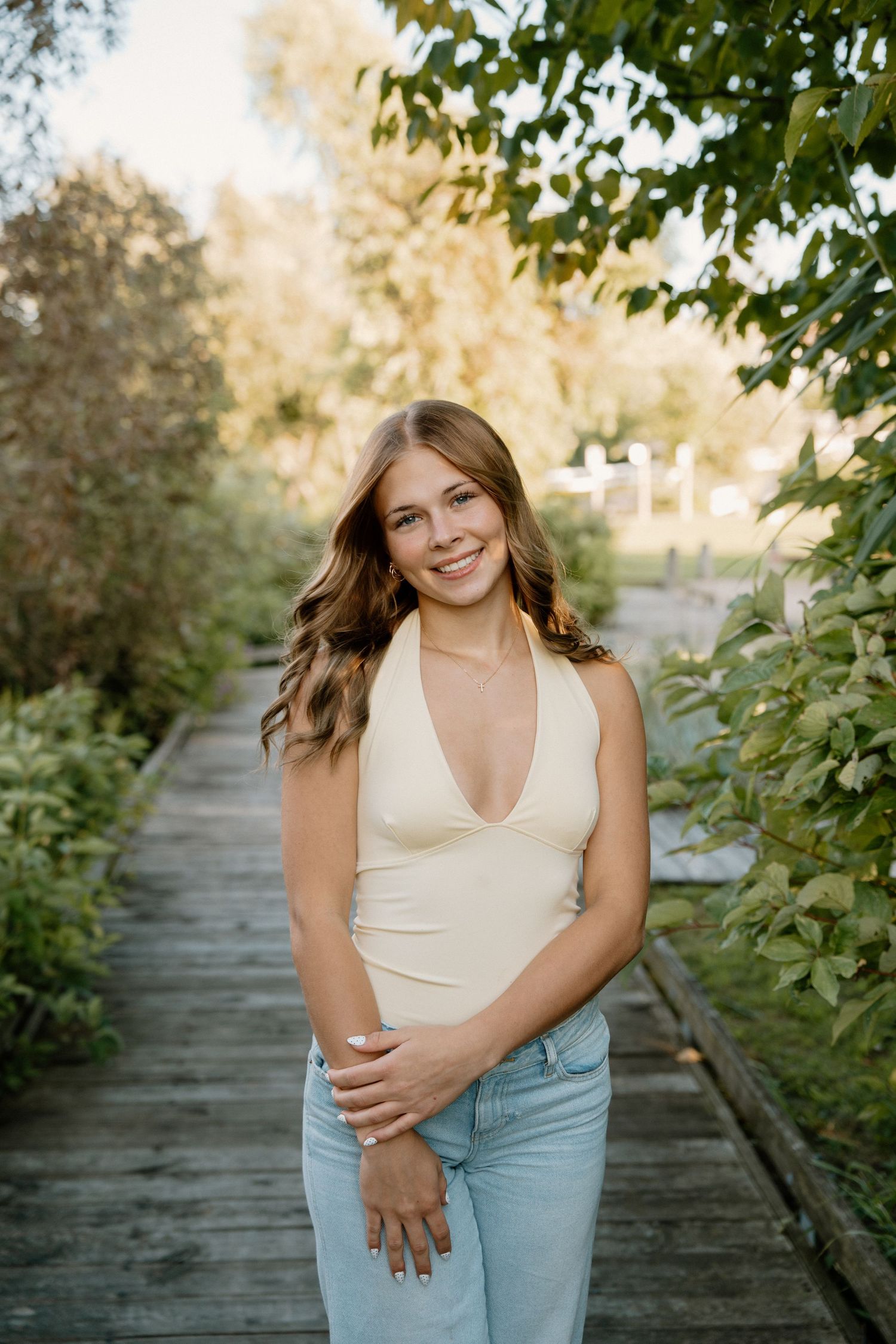 Young woman in cream halter top and jeans smiling on a wooden boardwalk surrounded by lush green foliage.
